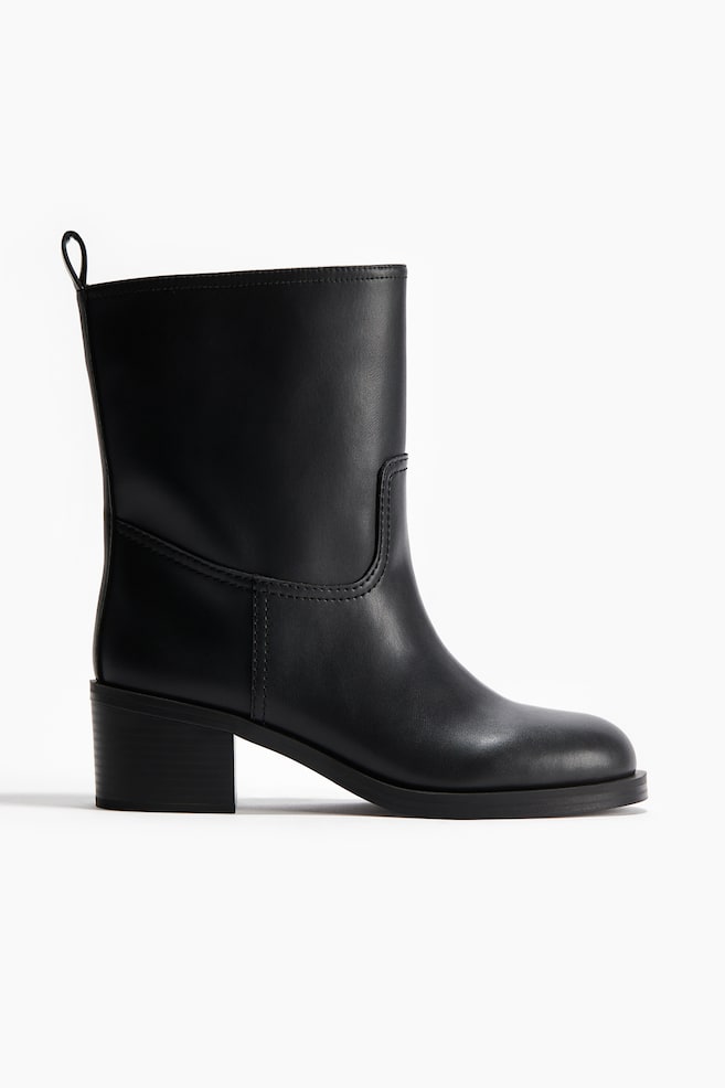 A black boot is presenting from a side angle, featuring a smooth finish and a mid-height shaft. It is detailed with U-shaped stitching on the side of the shaft, a rounded toe, a low block heel, and a small pull tab at the top back. - 1
