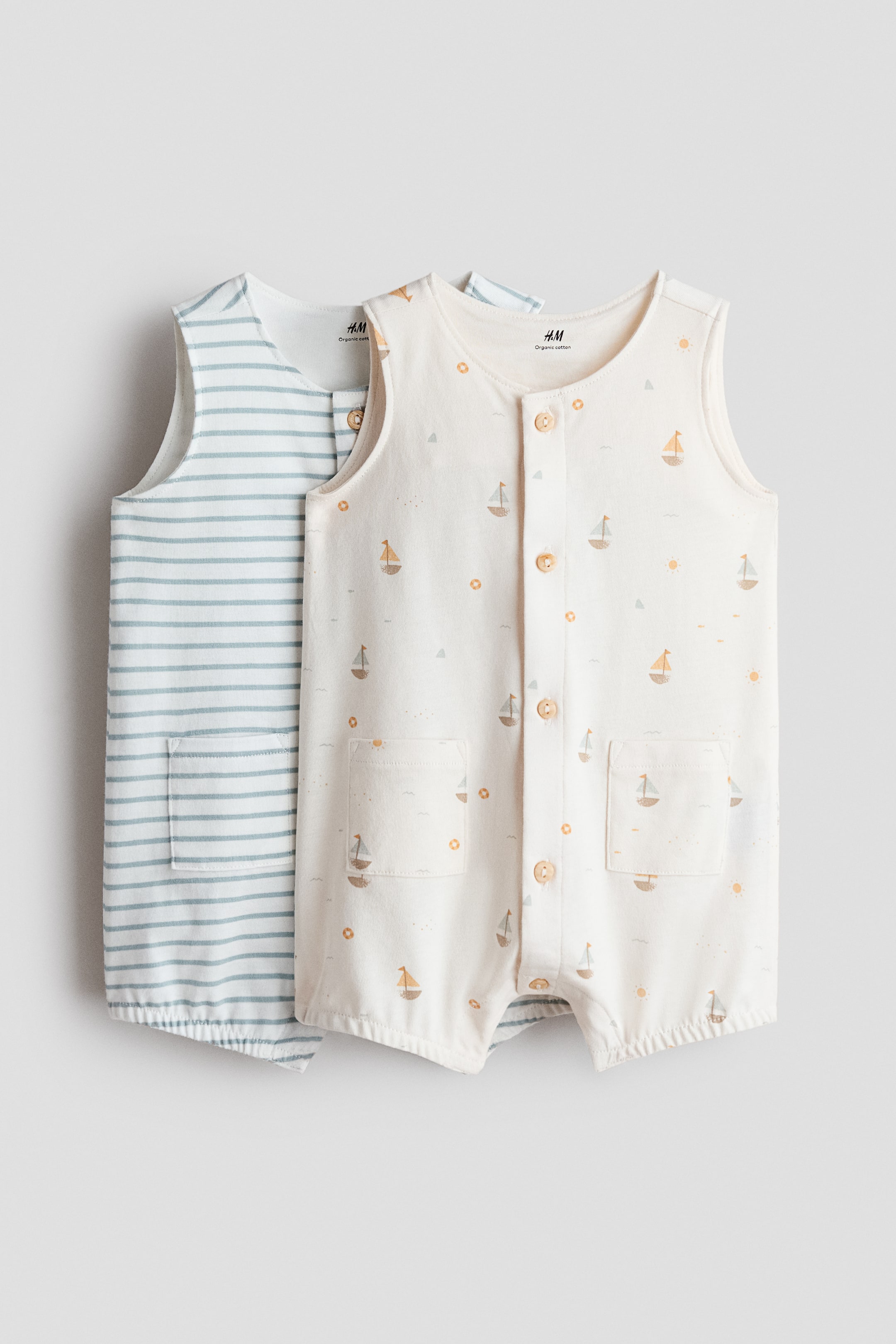 Ver imagen más grande: Se exhiben dos peleles para bebé, ambos con un diseño sin mangas, cierres frontales de botones y aberturas elásticas en las piernas. El pelele delantero es de color blanco roto, con un estampado de veleros y soles, y está diseñado con dos bolsillos de parche. El pelele situado detrás es blanco con rayas horizontales de color azul claro e incluye un bolsillo de parche.