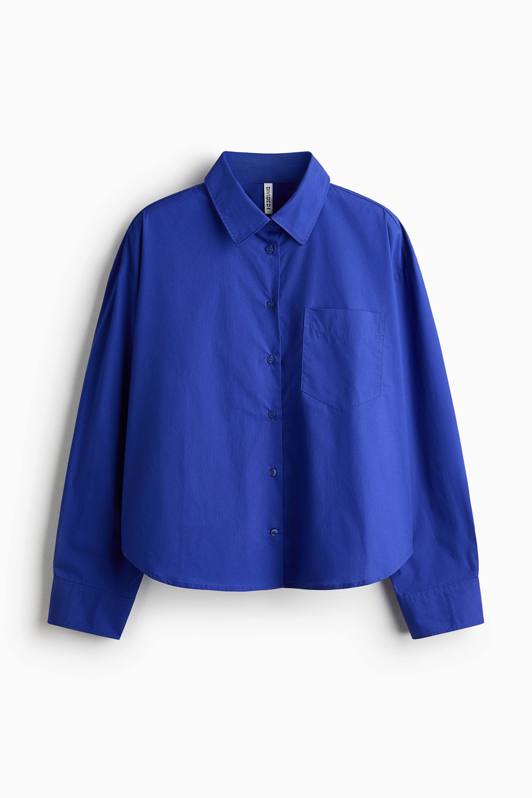 View larger image: The oversized blue shirt, facing front, features a crisp cotton poplin fabric, a collar, buttons down the front, and an open chest pocket. It also highlights dropped shoulders, long sleeves with buttoned cuffs, and a straight-cut hem with rounded side vents.