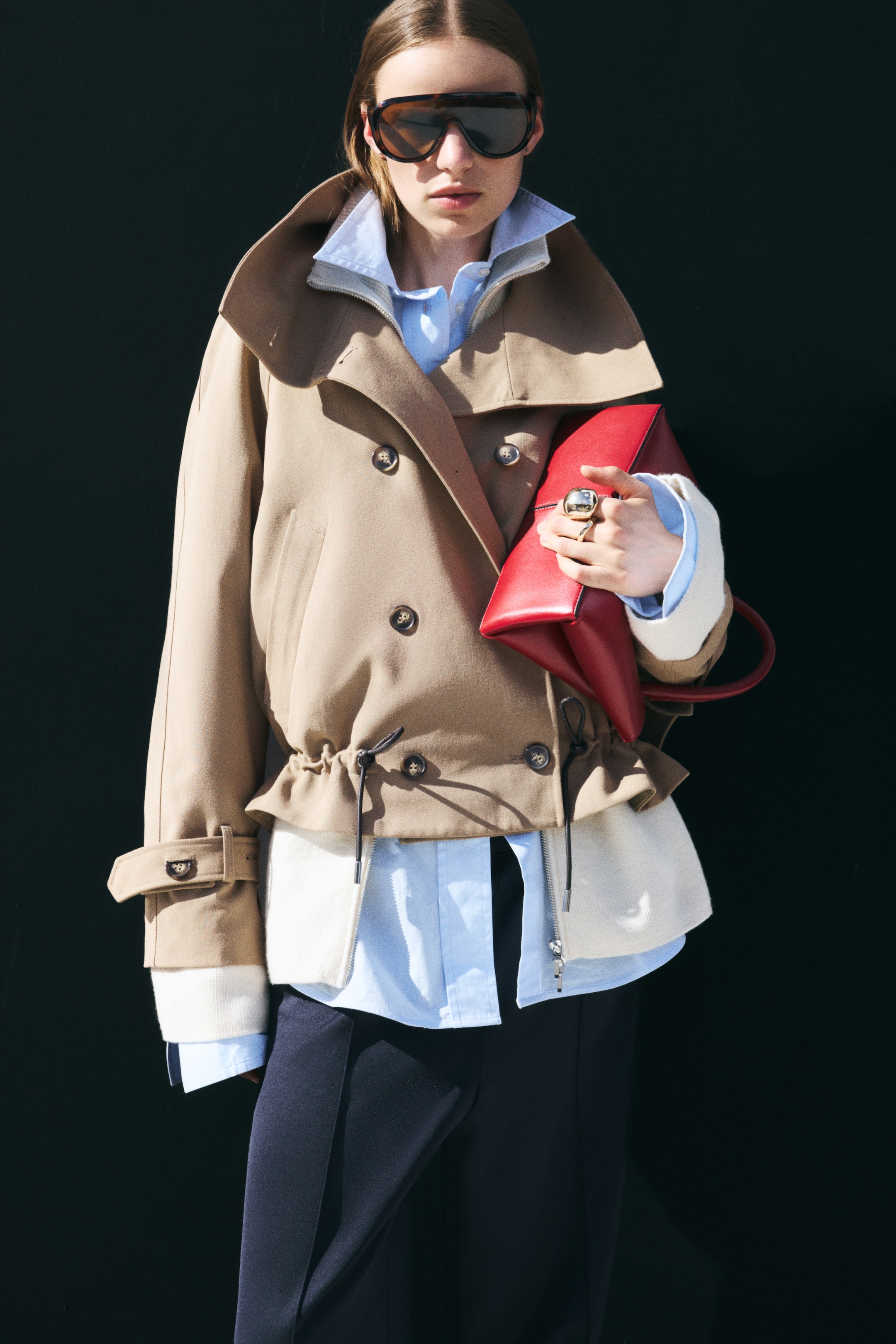 Visualizza l'immagine ingrandita: Un trench coat corto beige in twill, doppiopetto, indossato dalla modella, con ampio collo a imbuto, bottoni frontali e fondo arricciato con coulisse. Il cappotto è portato sopra una camicia azzurra con colletto e un top crema con zip, mentre sotto un braccio viene tenuta una borsa in pelle rossa.
