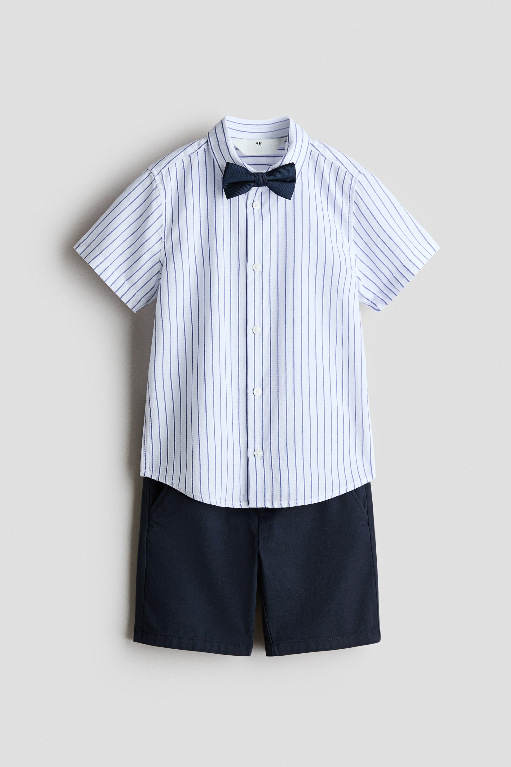 View larger image: A children's clothing set is showcasing a short-sleeved shirt, dark blue shorts, and a dark blue pre-tied bow tie, presented from the front. The white shirt features thin blue vertical stripes and buttons down the front, while the dark blue shorts display diagonal side pockets.