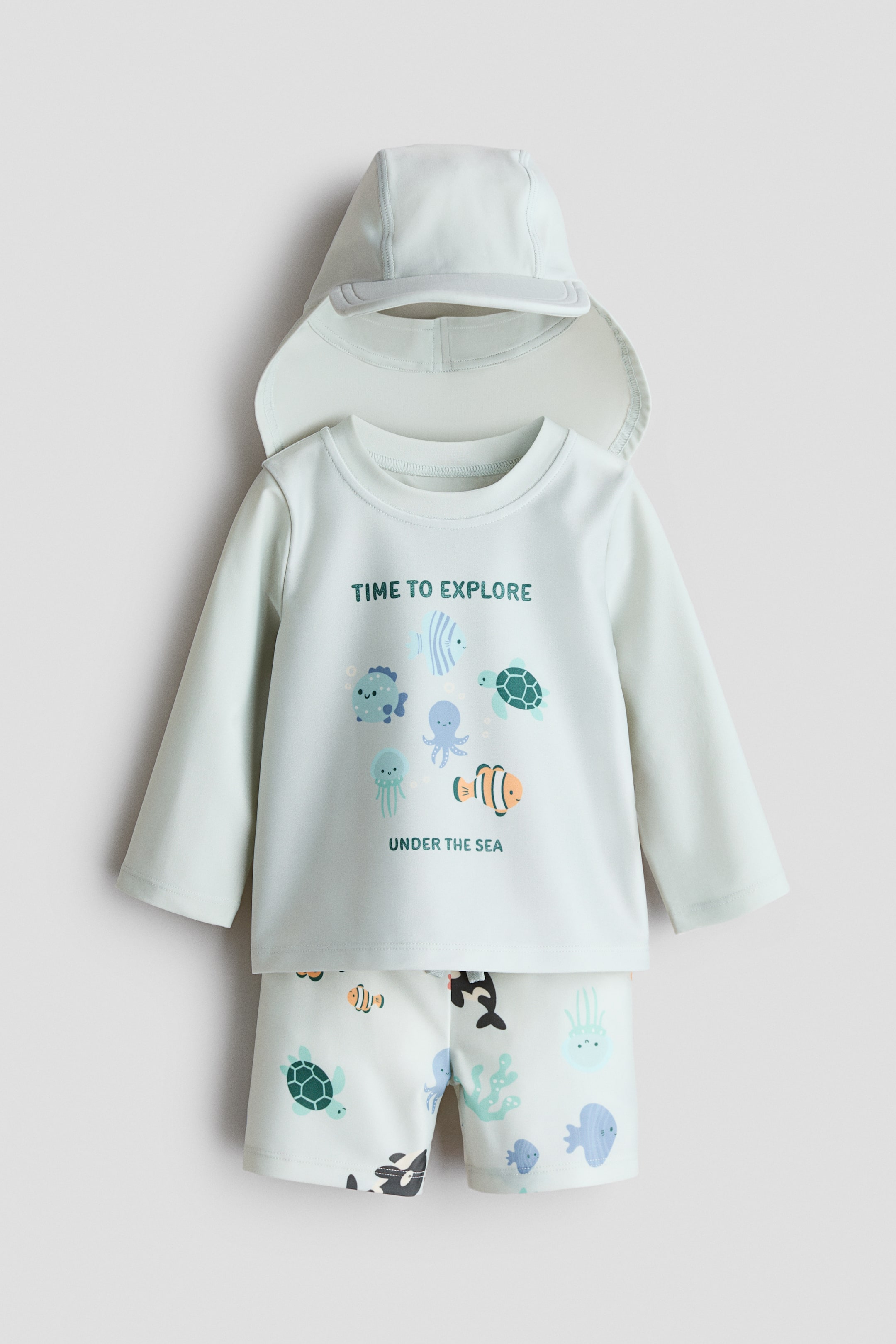 Agrandir l'image: Ensemble de bain trois pièces, présenté de face, comprenant un haut de maillot de bain vert menthe clair avec un col rond et des manches longues raglan, décoré d'illustrations colorées de créatures marines et de texte. Un short de bain assorti avec une taille élastiquée présente un imprimé intégral de créatures marines, et une casquette coordonnée avec une visière à l'avant et un rabat long pour le cou repose au-dessus du haut.