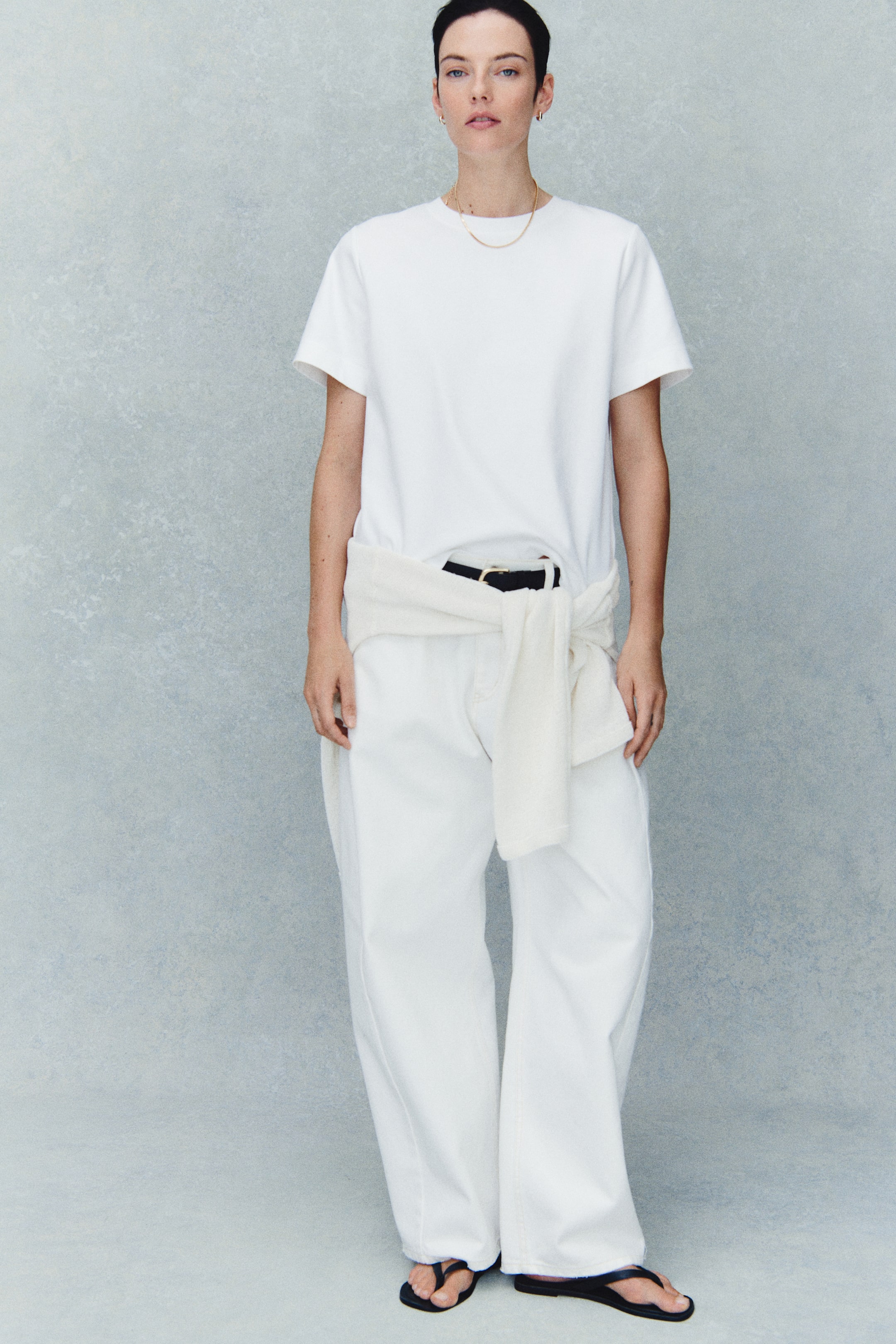 View larger image: The model is wearing a white, loose-fitting T-shirt with a crew neckline and dropped shoulders. It’s tucked into white wide-leg trousers, styled with a black belt and a cream sweater tied around the waist.
