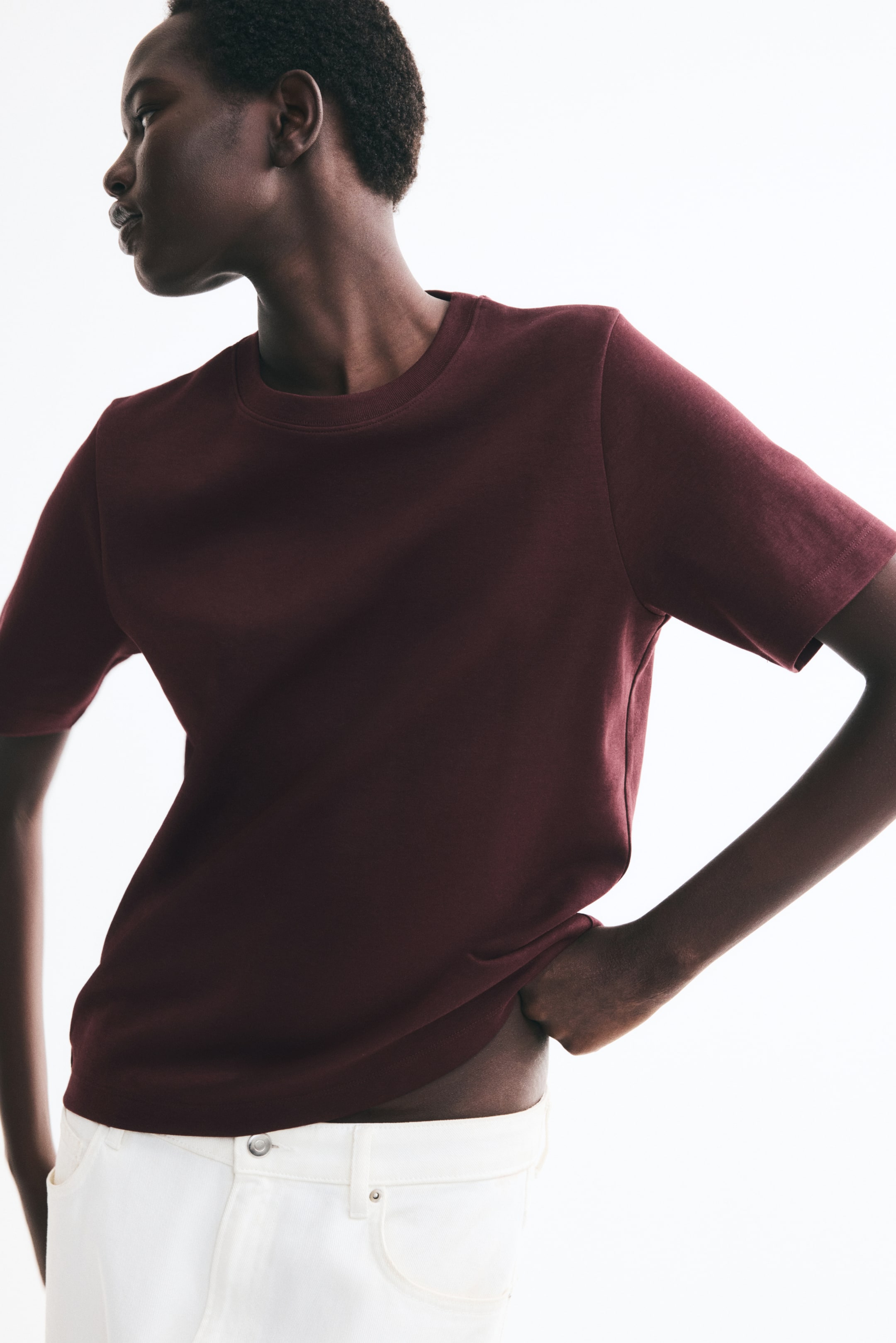 Visualizza l'immagine ingrandita: Una t-shirt bordeaux in jersey interlock pesante è indossata dalla modella, con scollo tondo rifinito a costina e maniche corte. La modella solleva leggermente l’orlo dritto all’altezza dell’anca, abbinando la t-shirt a pantaloni bianchi.