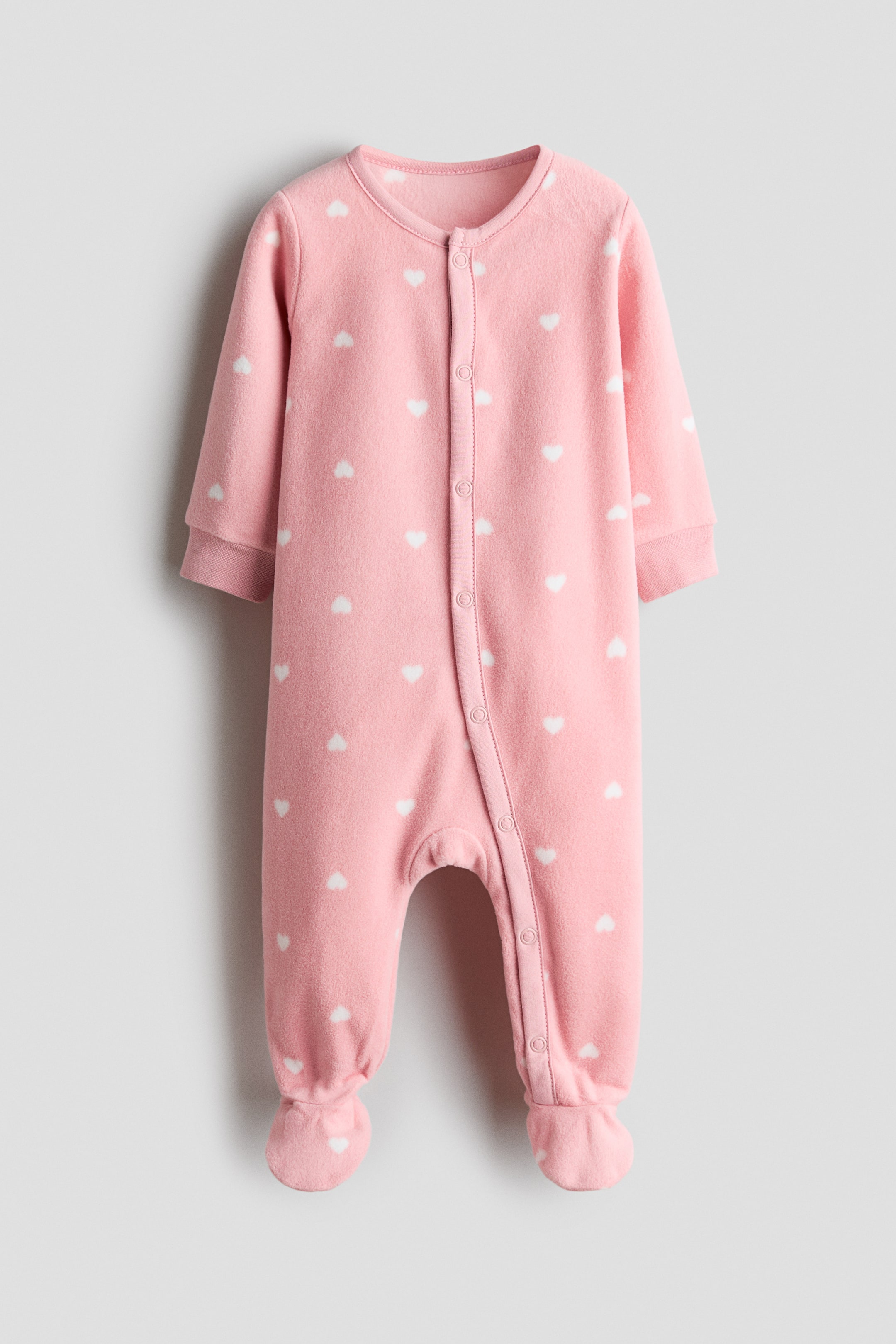 Größeres Bild ansehen: Ein rosafarbener Fleece-Schlafanzug für Babys mit einem Muster aus weißen Herzen. Er ist mit Rundhalsausschnitt, langen Ärmeln mit Rippbündchen und integrierten Füßchen gestaltet und verfügt über einen Druckknopfverschluss, der sich vorne und entlang der Innenseite der Beine erstreckt.