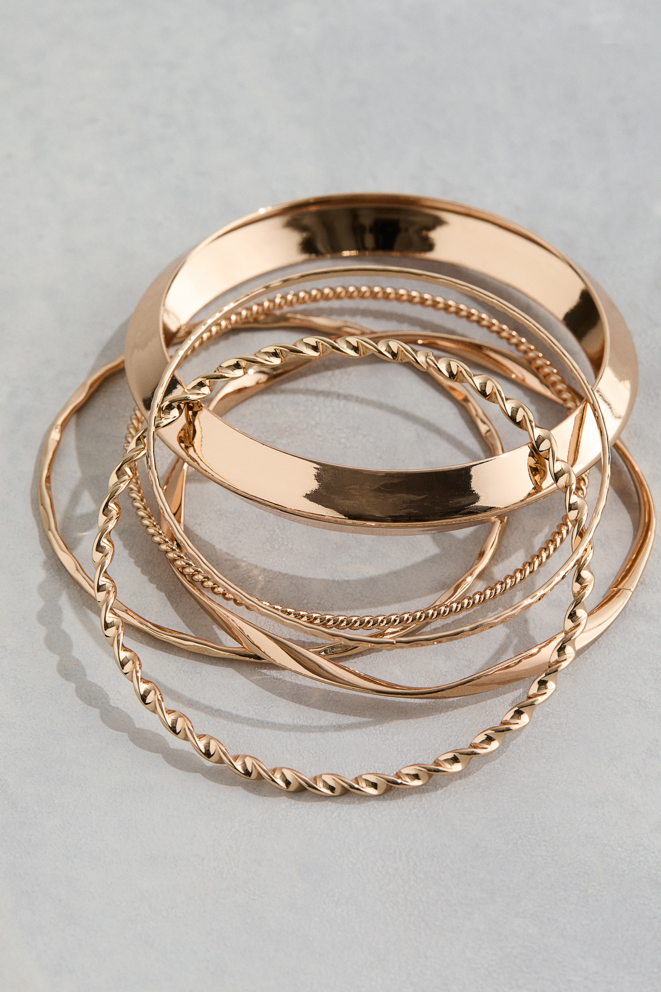 View larger image: "A collection of gold-toned bangles is featuring diverse designs, including broad polished bangles, twisted bangles, a beaded bangle, and a slender hammered bangle. The bangles are overlapping and resting together."