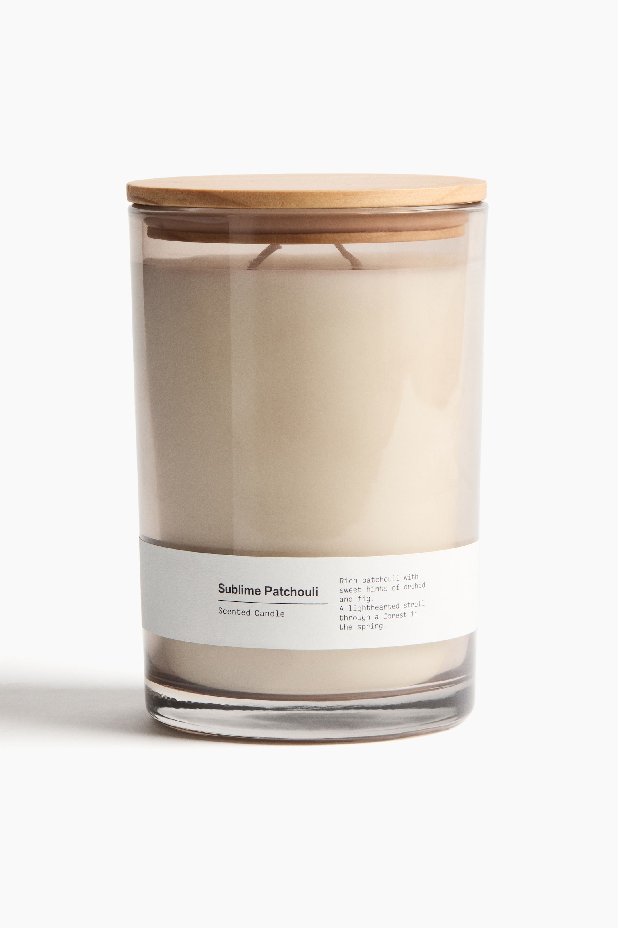 View larger image: A scented candle is featured FRONT in a clear cylindrical glass jar, filled with light beige wax and a visible wick. A light-colored circular wooden lid is resting on the top, and a white rectangular label is wrapped around the front, displaying "Sublime Patchouli Scented Candle" and a detailed scent description.