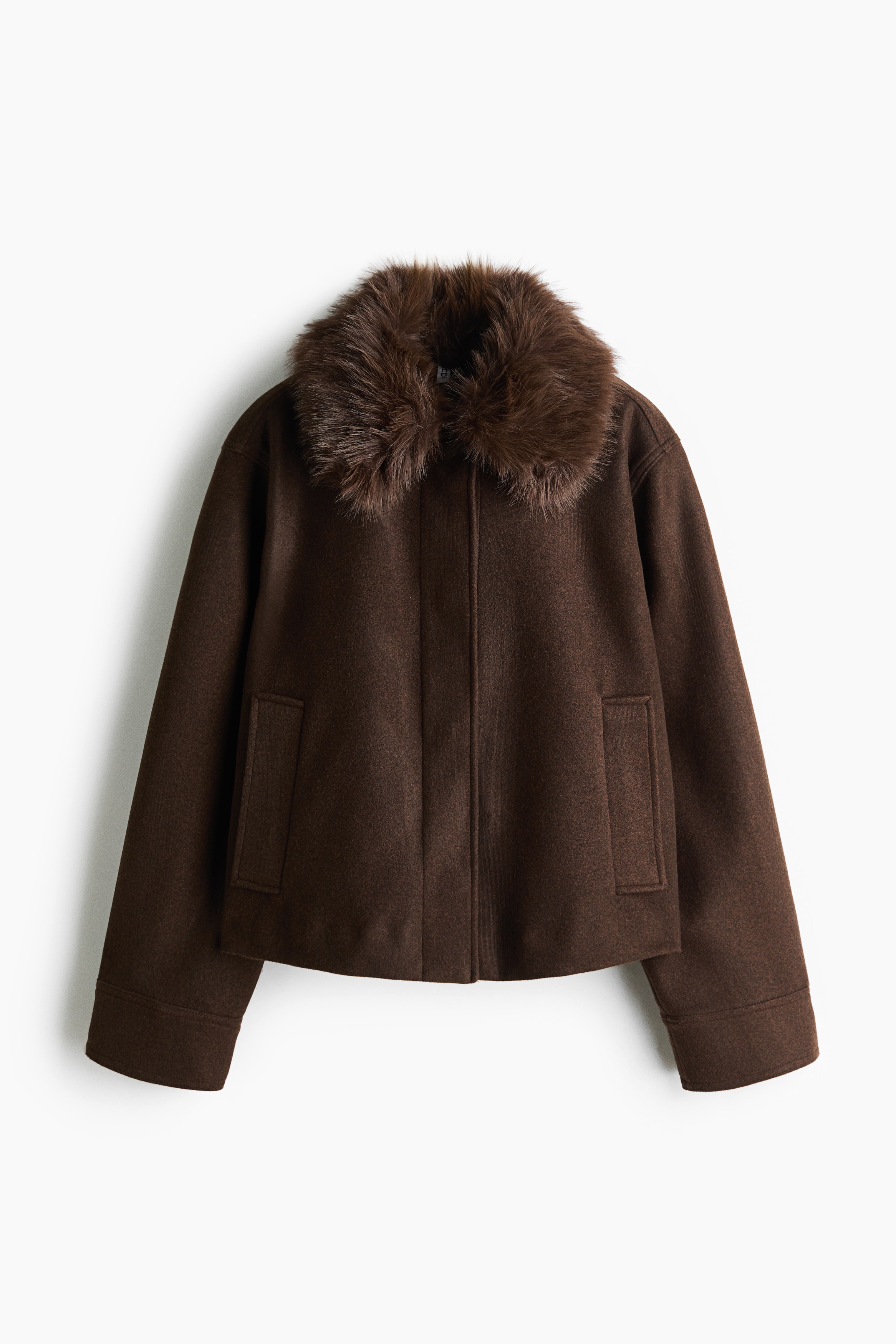 A short, dark brown jacket in a felted fabric is being worn by the model, who is standing with one hand tucked into the front welt pocket. The jacket features a brown fluffy faux-fur collar and is layered over a dark brown knit top and a dark grey skirt.