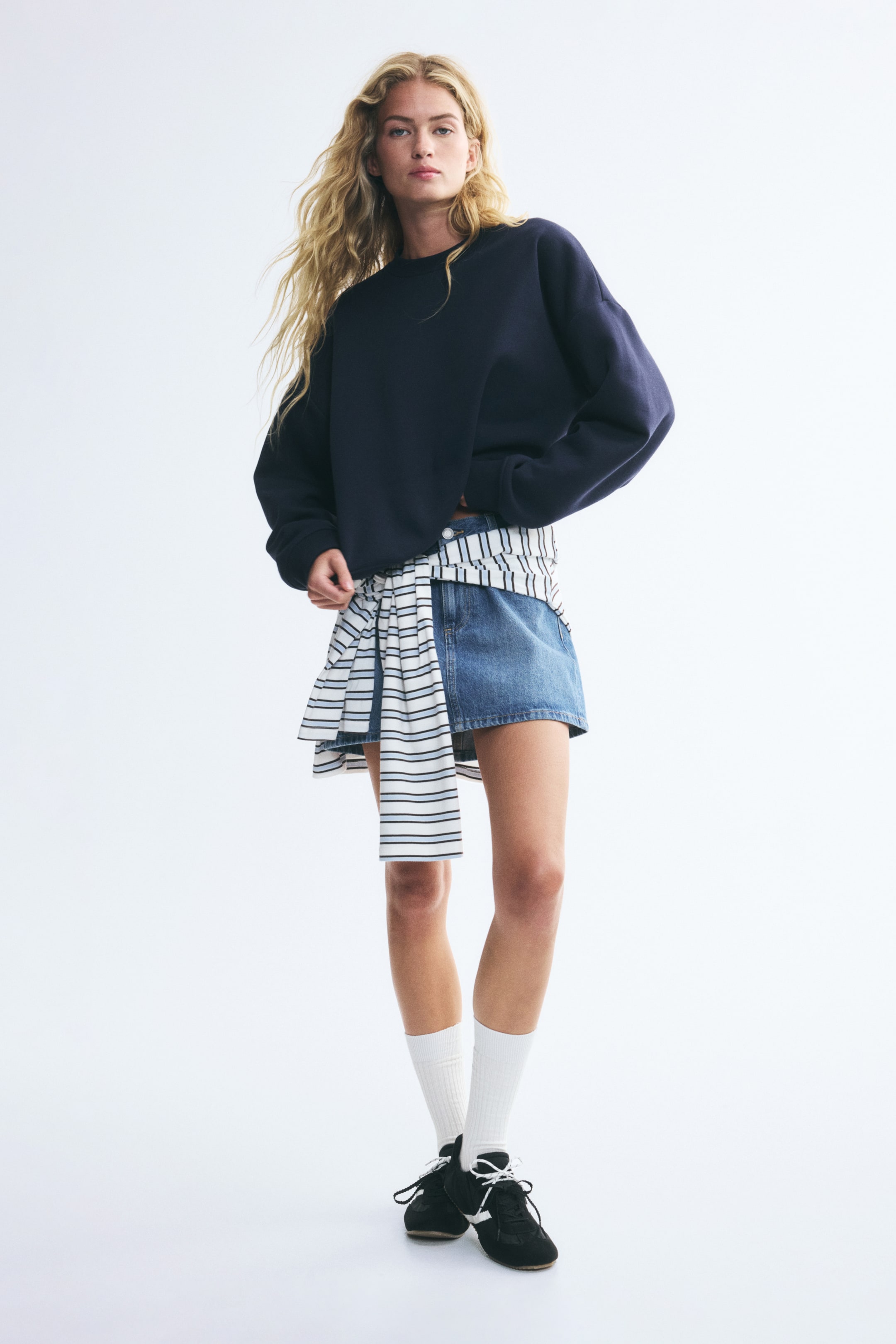 View larger image: An oversized navy blue sweatshirt featuring a round neckline, dropped shoulders, and ribbed cuffs is worn by the model. The sweatshirt is styled with a blue denim mini skirt, a striped shirt tied around the waist, white socks, and black sneakers.