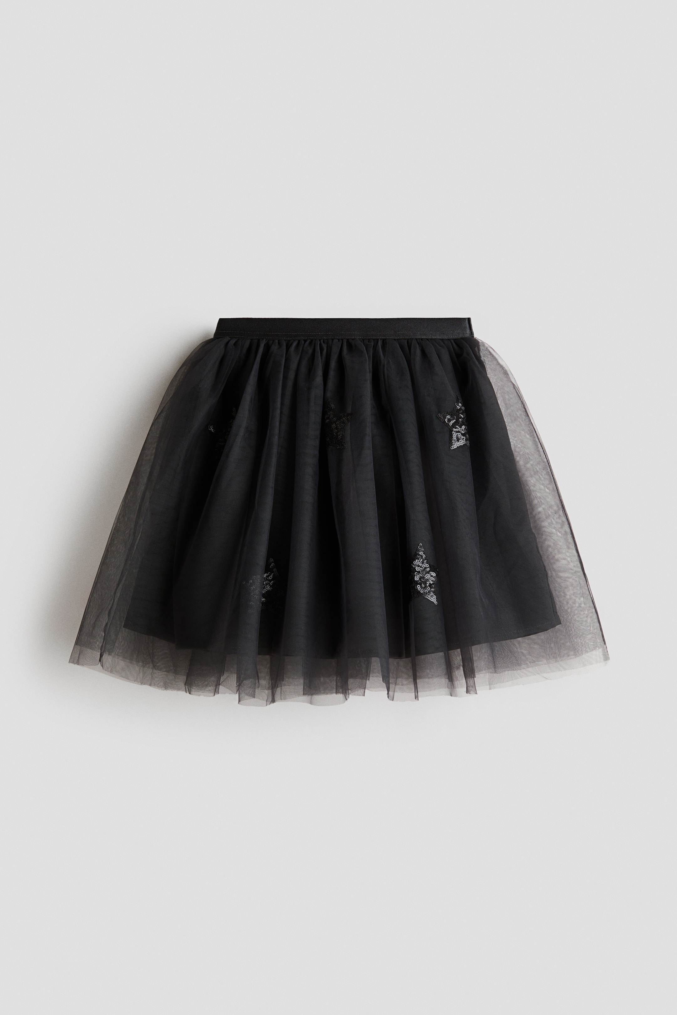 View larger image: A black flared tulle skirt is shown from the front, featuring an elasticated waistband with gathers that add volume to the flared silhouette. The sheer tulle outer layer is adorned with scattered shiny sequin star motifs and layered over a solid black lining.