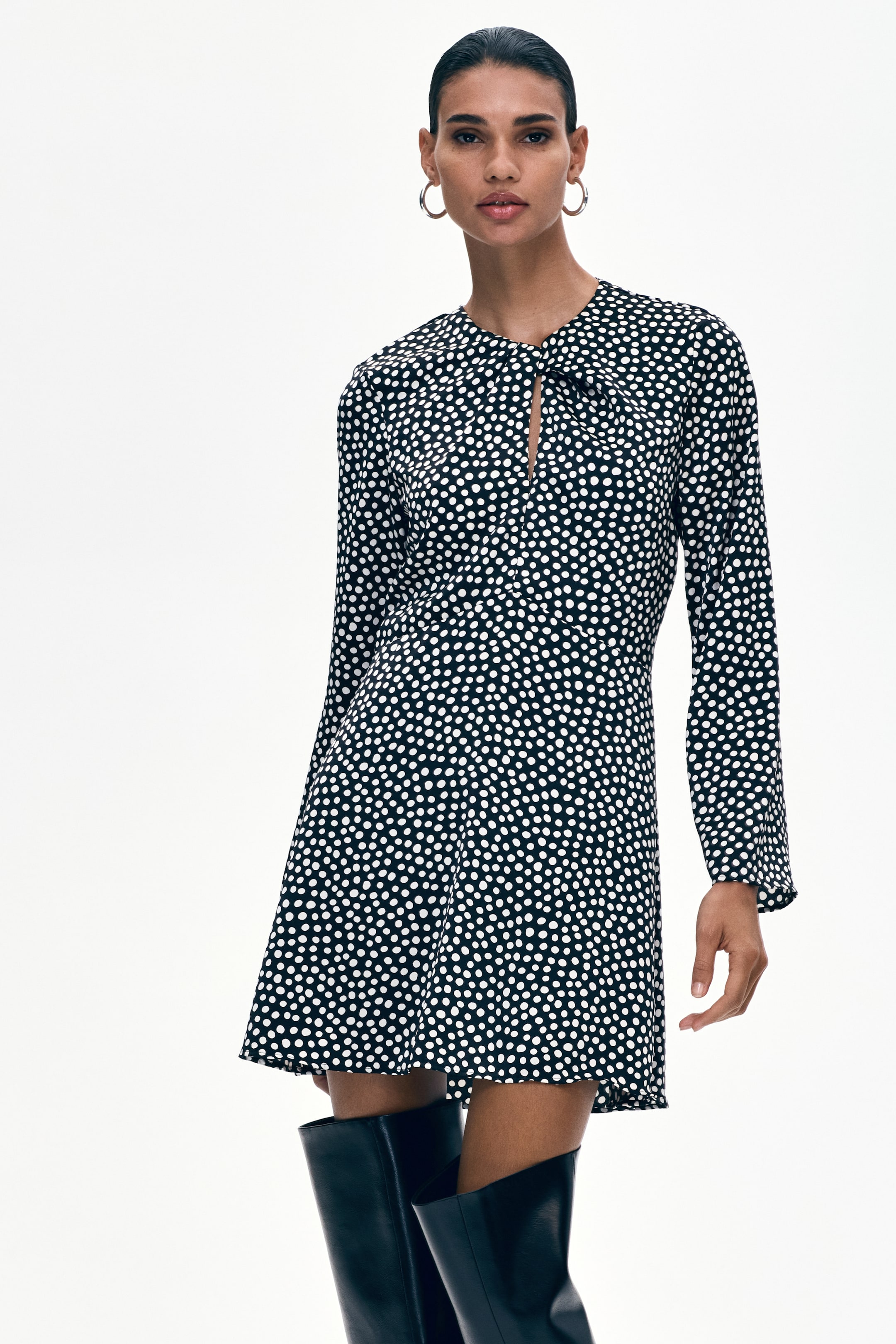 View larger image: A black dress with white polka dots features a round neckline with a keyhole cut-out and a twisted fabric detail at the front. It is styled with long sleeves, a fitted waist, and a flared skirt ending above the knee, while facing forward.