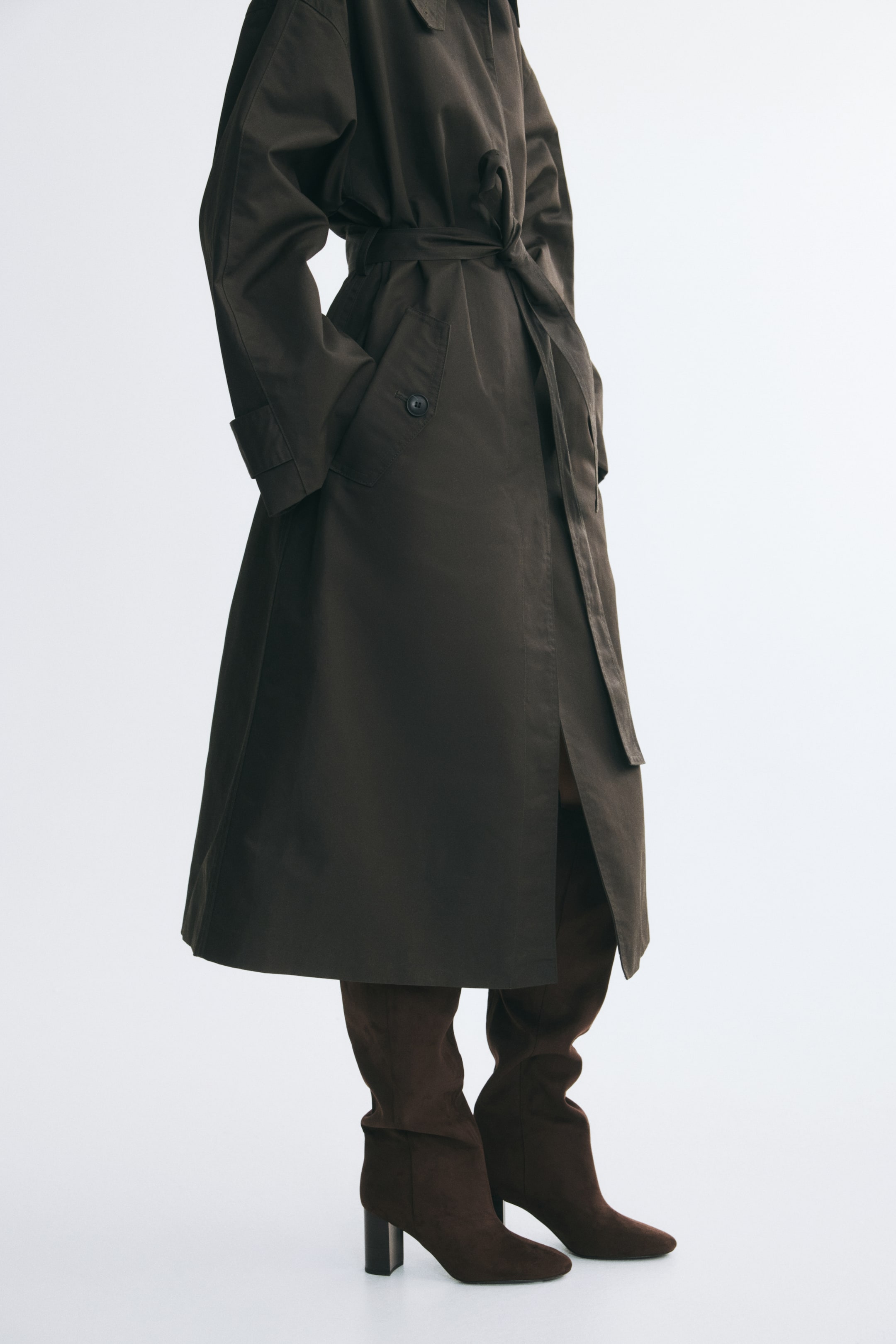 Größeres Bild ansehen: Dunkelbraune Stiefel mit Blockabsatz, weicher angerauter Oberfläche und mandelförmiger Spitze werden vom Model getragen. Die kniehohen Schäfte der Stiefel sind unter einem dunkelbraunen Trenchcoat mit Gürtel getragen.