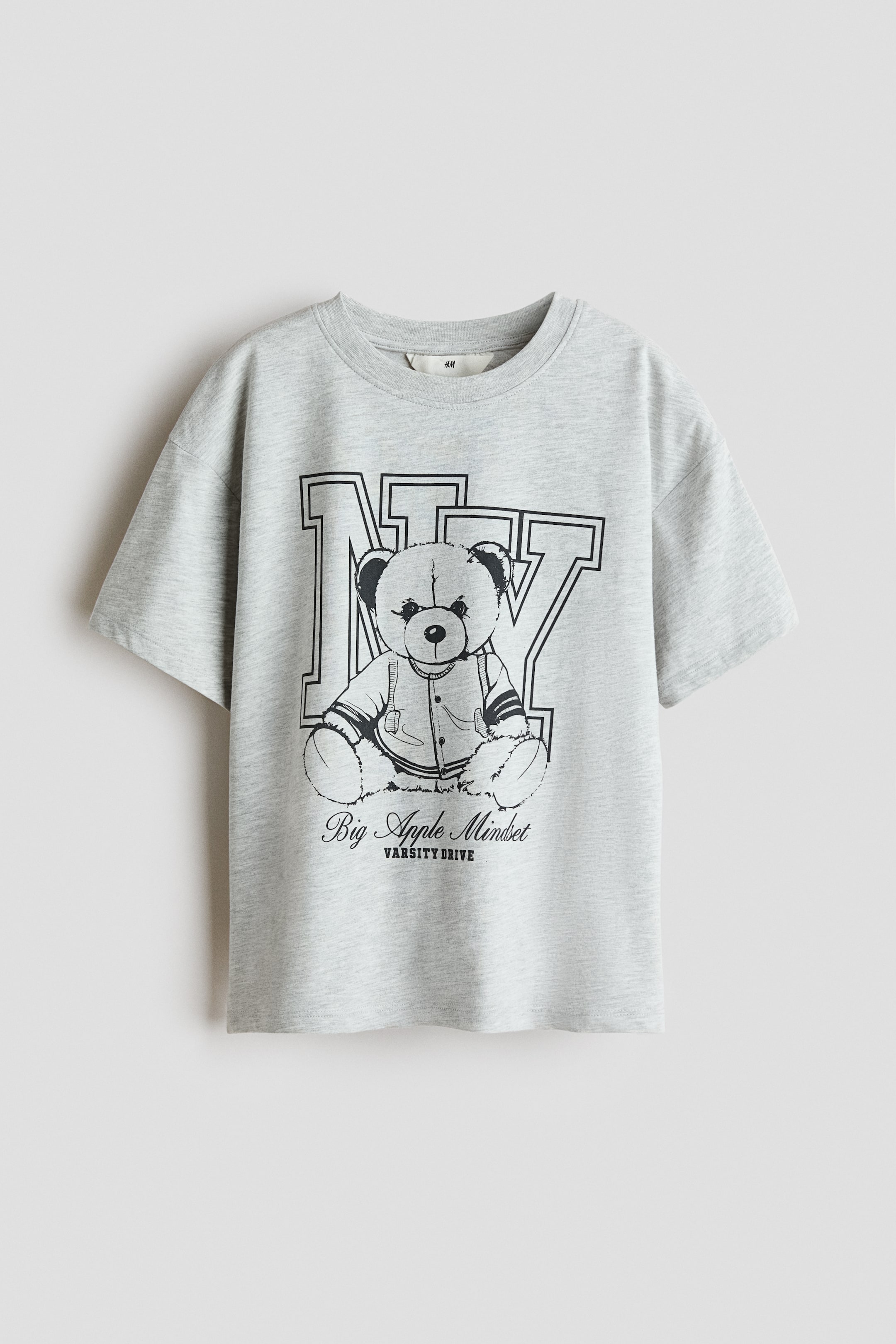 Agrandir l'image: Un t-shirt en jersey de coton gris clair chiné surdimensionné est présenté de face, avec un col rond bordé de côtes, des épaules tombantes et un ourlet droit. Un grand imprimé graphique noir recouvre le devant, représentant des lettres stylisées "NY" avec un ours en peluche portant une veste universitaire, et le texte "Big Apple Mindset VARSITY DRIVE" en dessous.