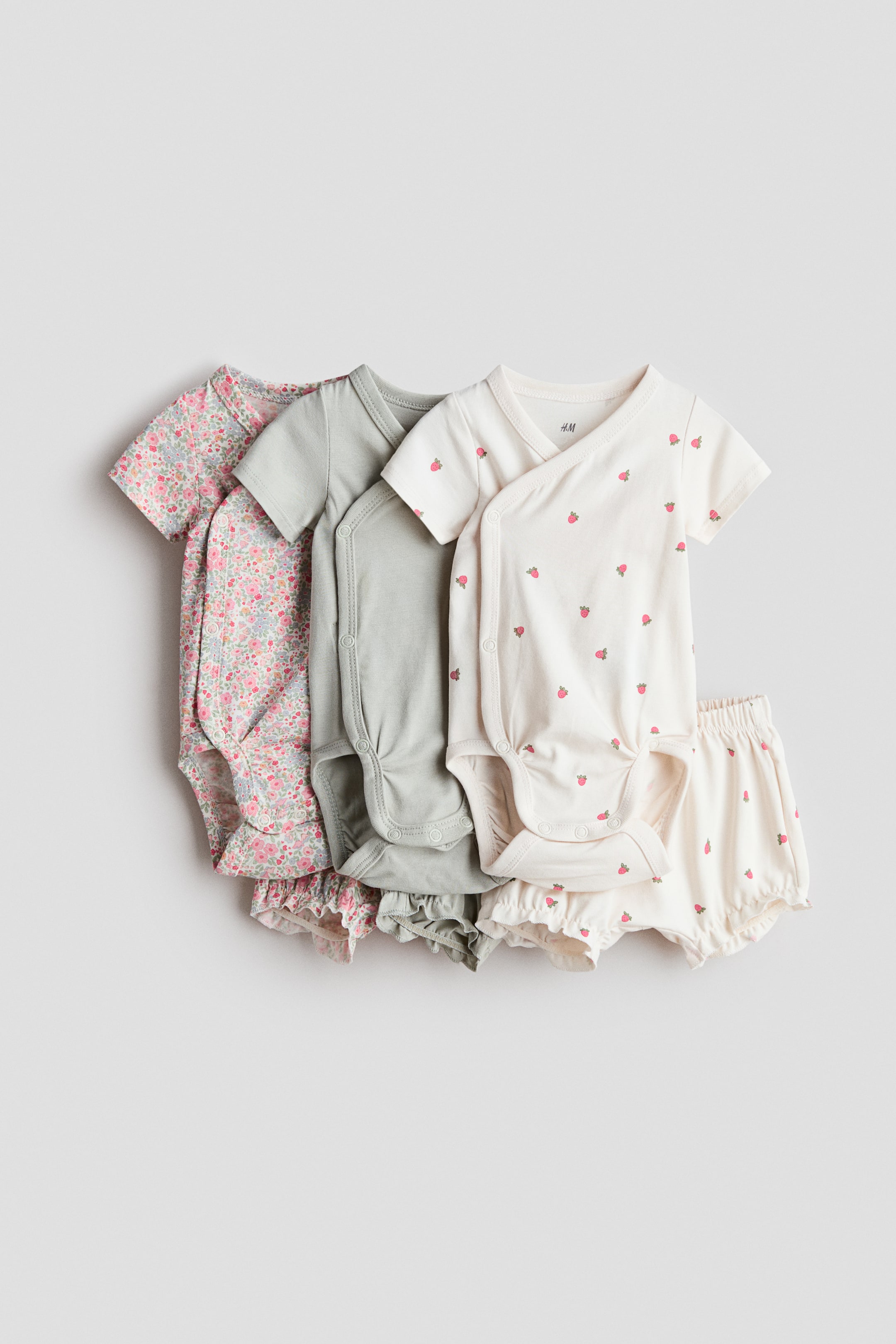 View larger image: Presenting a set of three short-sleeved cotton jersey wrapover bodysuits paired with three matching shorts. The bodysuits feature wrapover fronts with snap closures at the sides and crotch, available in a pink floral print, solid sage green, and cream with a strawberry pattern. Each bodysuit is complemented by coordinating shorts, designed with elasticated waists for comfort.