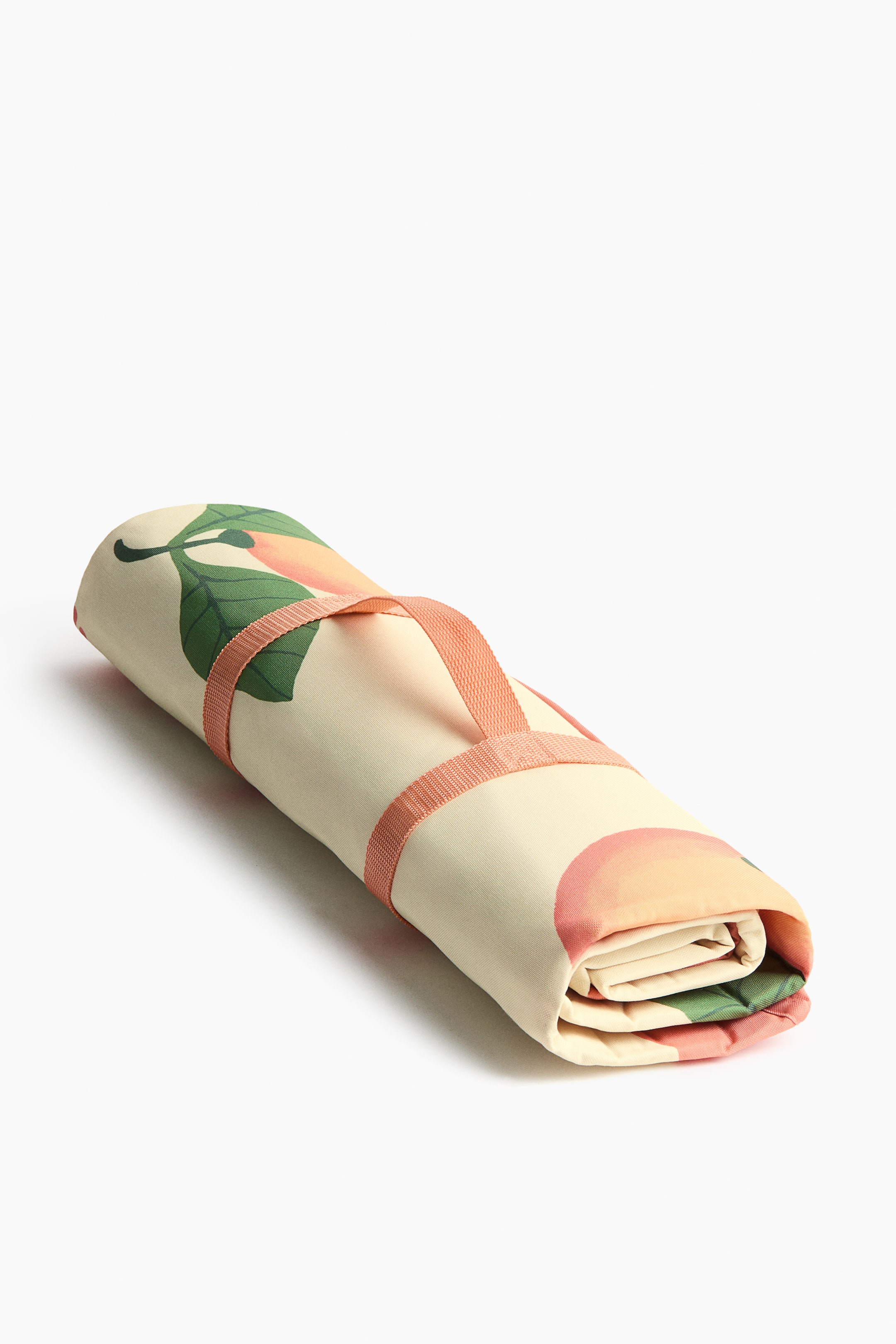 Ampliar la imagen: Una manta de picnic está enrollada horizontalmente, con un fondo crema y un patrón de hojas en tonos durazno y verde. Dos correas de tela rosa la aseguran, formando un asa en el centro.