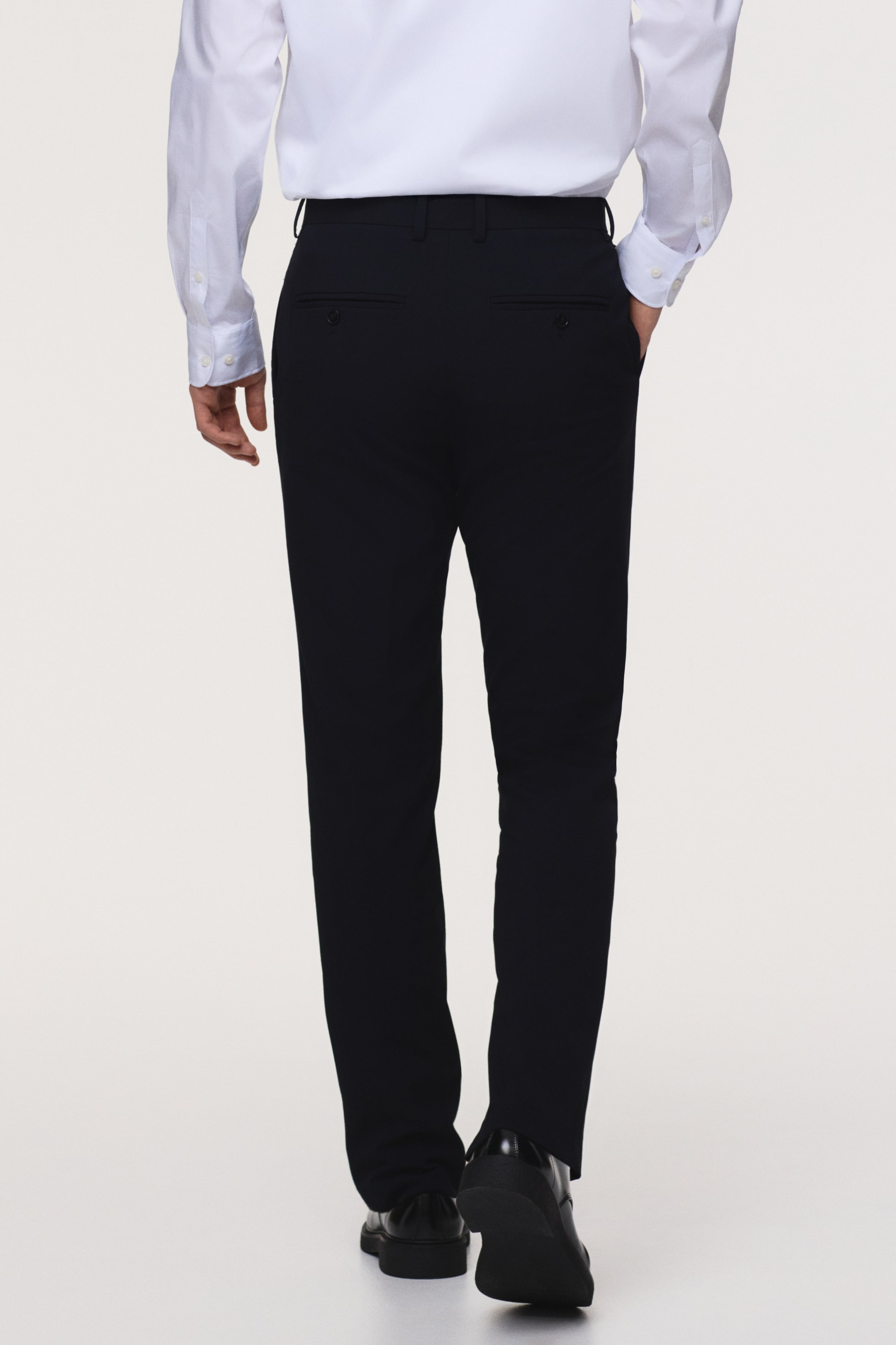 Agrandir l'image: Le modèle porte un pantalon sombre ajusté avec une coupe slim, doté de poches arrière passepoilées avec boutons et de plis sur les jambes. Le modèle se tient debout, une main dans une poche latérale, et le pantalon est assorti d'une chemise blanche et de chaussures habillées noires.
