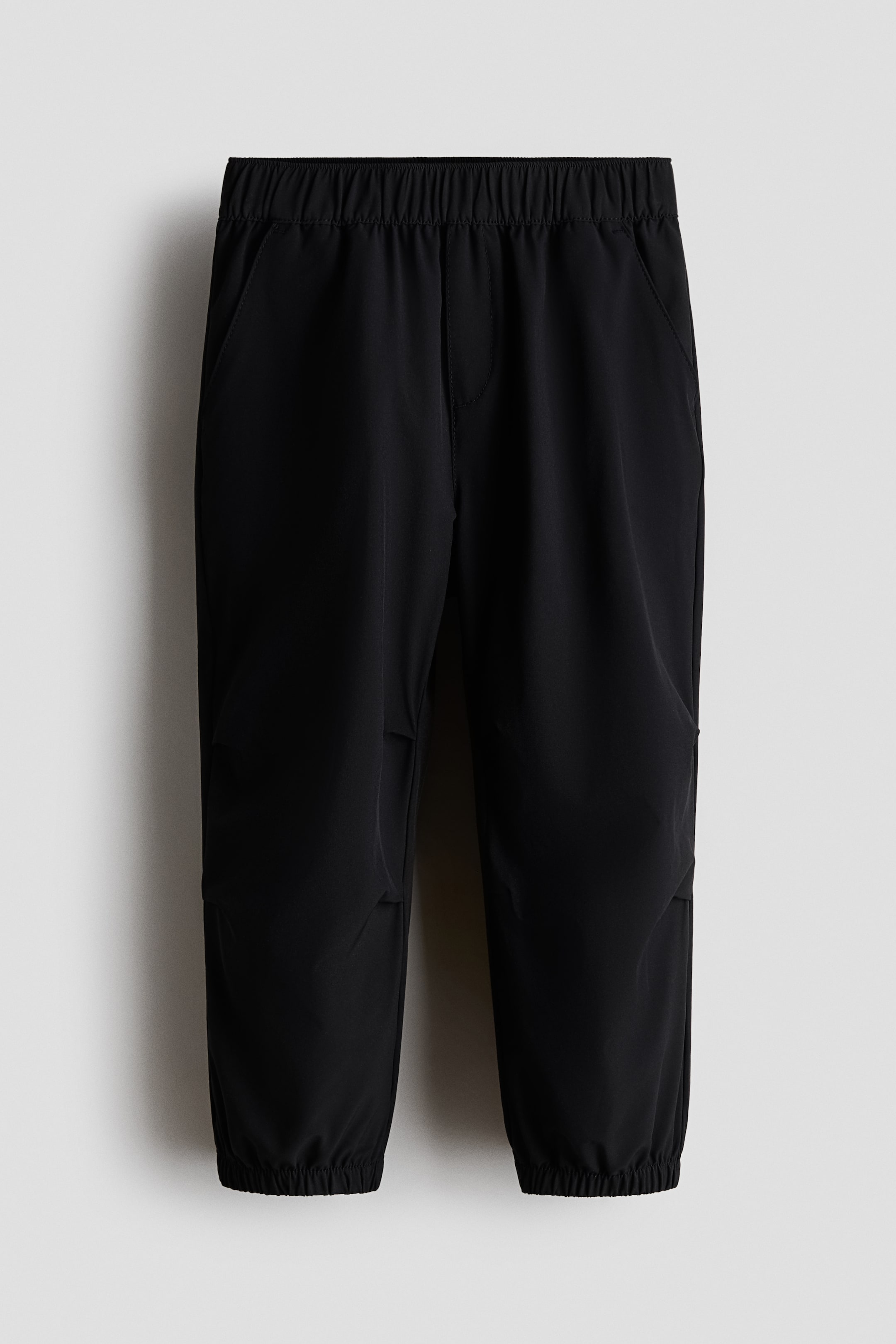 拡大画像を表示: ブラックジョガーパンツは、ウエストにゴムが入っており、前面には斜めのサイドポケットが付いています。また、フェイクフライ、膝のダーツ、裾のカバー付きゴムも特徴です。