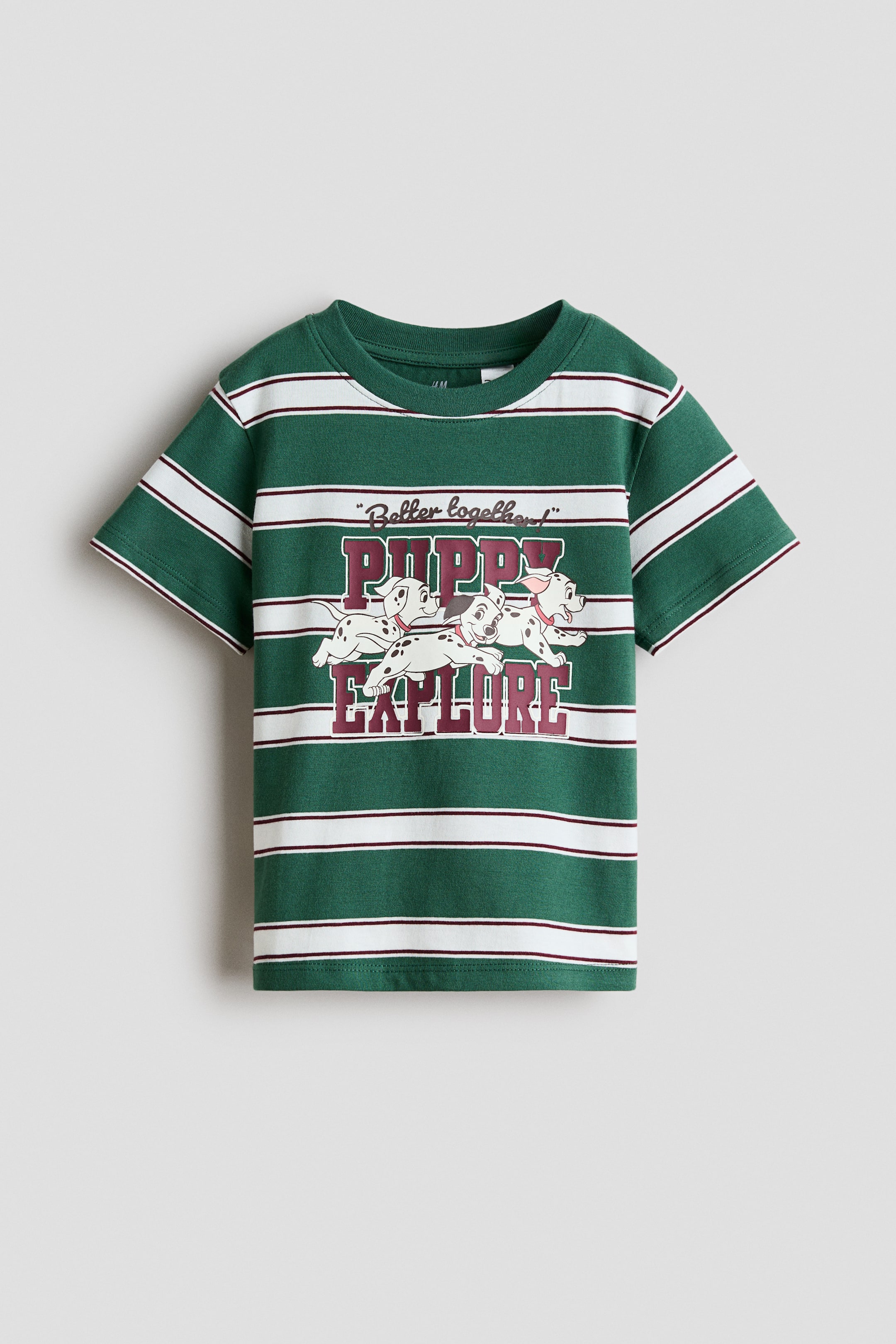 View larger image: A green short-sleeved t-shirt is lying flat, featuring horizontal white stripes bordered by thin maroon lines. It is displaying a graphic print on the front, showing three Dalmatian puppies positioned between the maroon and white collegiate-style text "PUPPY" and "EXPLORE," with "Better together" in a script font above.