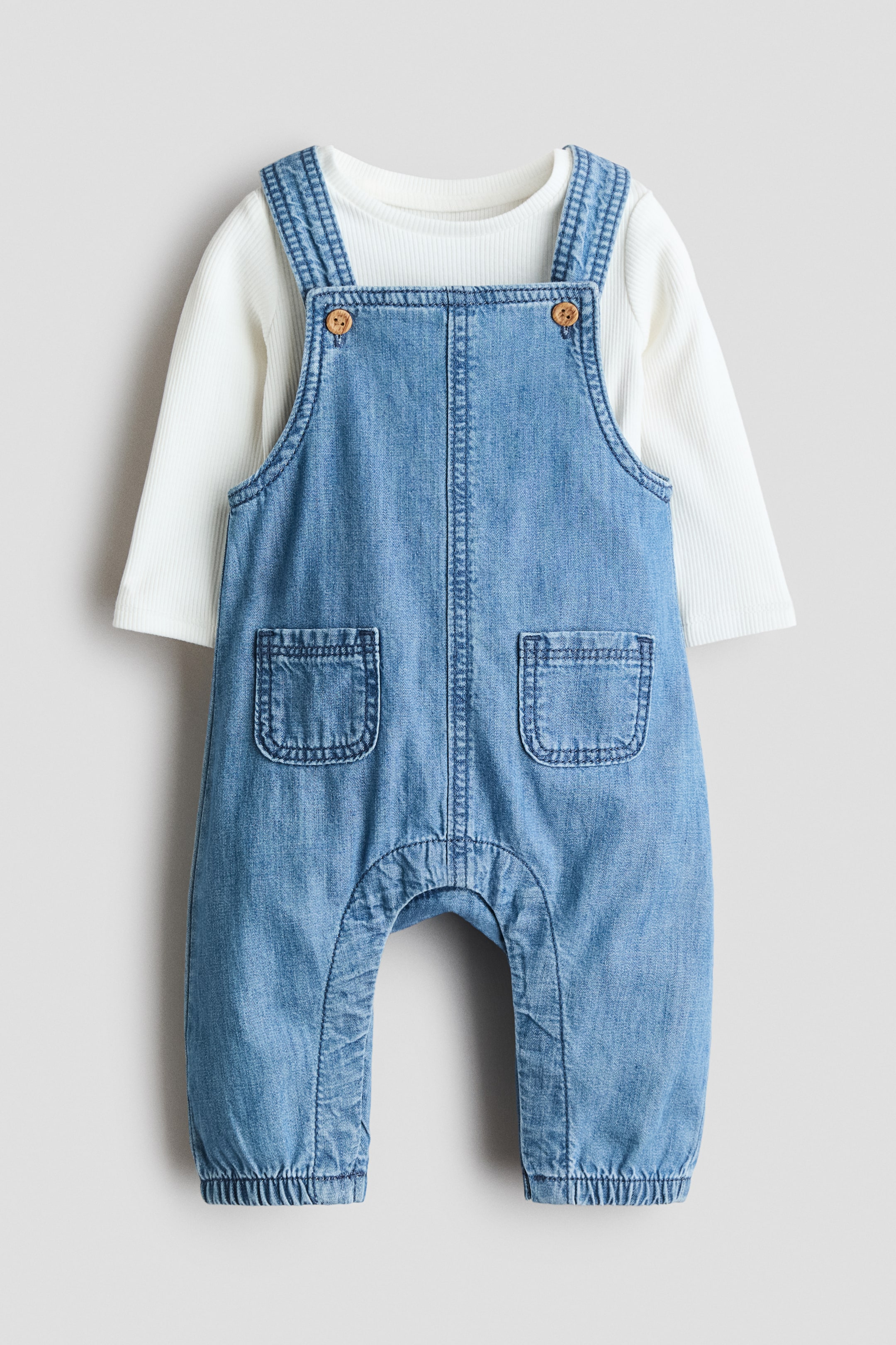 Grösseres Bild ansehen: Ein Babyset bestehend aus hellblauen Denim-Latzhosen, von vorne gesehen, über einem weißen gerippten Langarm-Body geschichtet. Die Latzhosen zeigen Schulterträger mit Holzoptik-Knopfverschlüssen, zwei offenen Fronttaschen und gerafften elastischen Säumen, während der runde Halsausschnitt und die Bündchen des Bodys sichtbar sind.