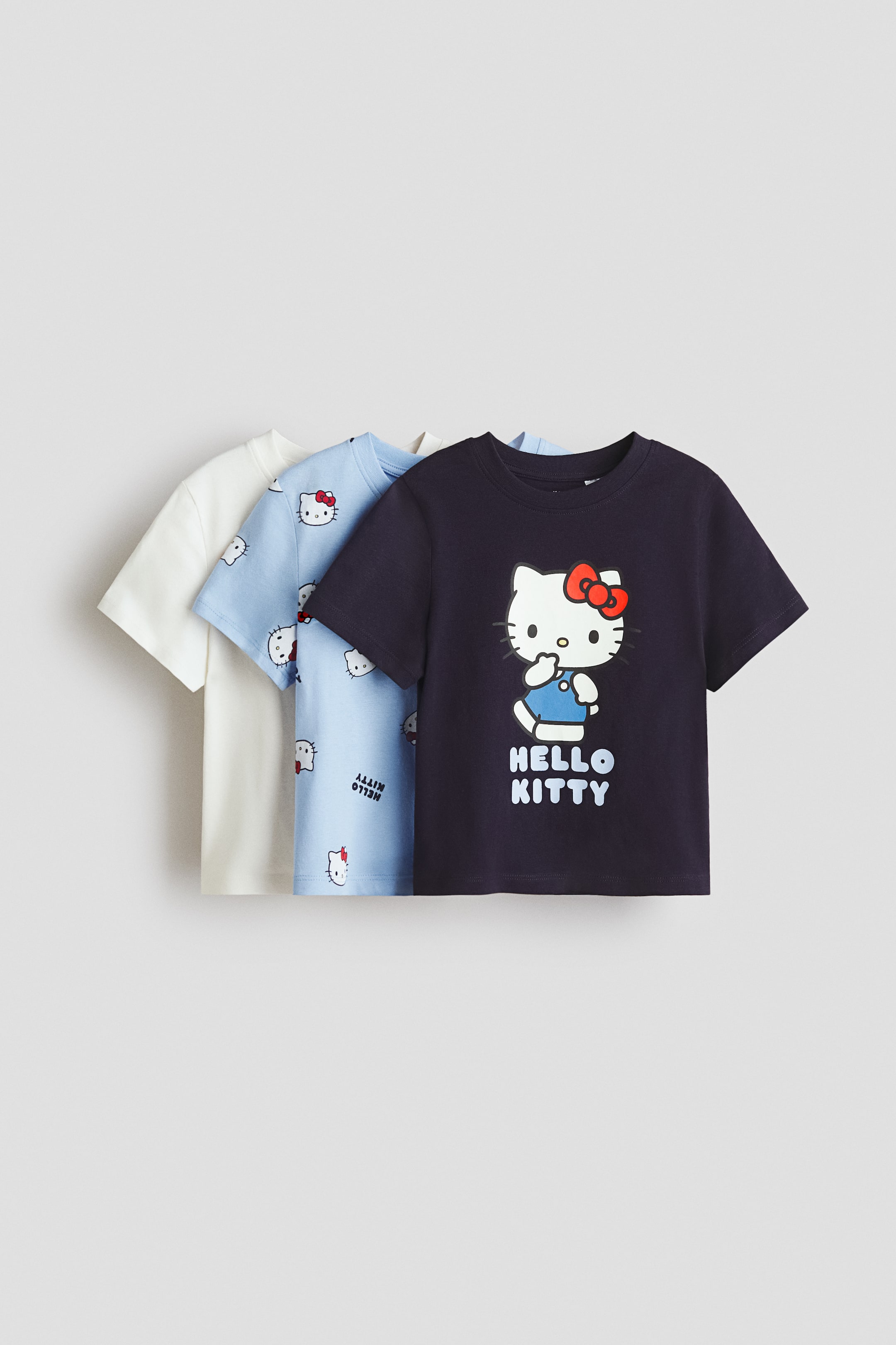 הצגת תמונה גדולה יותר: סט חולצות קצרות שרוול מבד ג'רזי כותנה רכה מוצגות בצד הקדמי שלהן, מסודרות בתצוגה שכבתית משמאל לימין בצבעי קרם, כחול בהיר וכחול כהה. החולצה הכחולה בהירה מציגה הדפס כולל של ראשים של הלו קיטי וטקסט הפוך "HELLO KITTY", והחולצה הכחולה כהה מציגה הדפס מרכזי גדול של הלו קיטי לובשת אוברול כחול עם טקסט "HELLO KITTY" מתחת.
