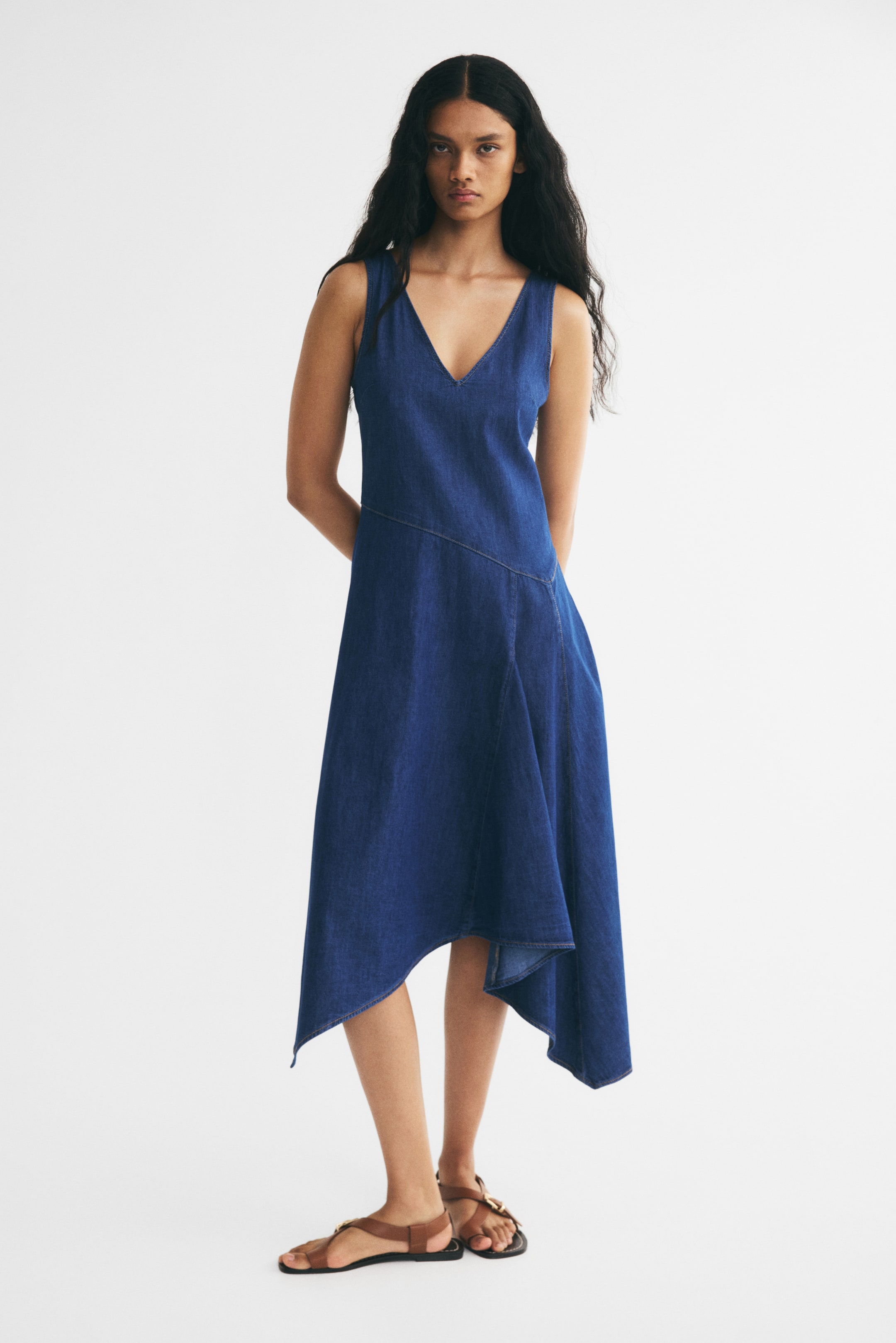 View larger image: A sleeveless, calf-length blue denim dress is being worn by the model, featuring a V-neck, a diagonal waist seam, and a flared skirt with a handkerchief hem. The dress is paired with flat brown leather sandals.