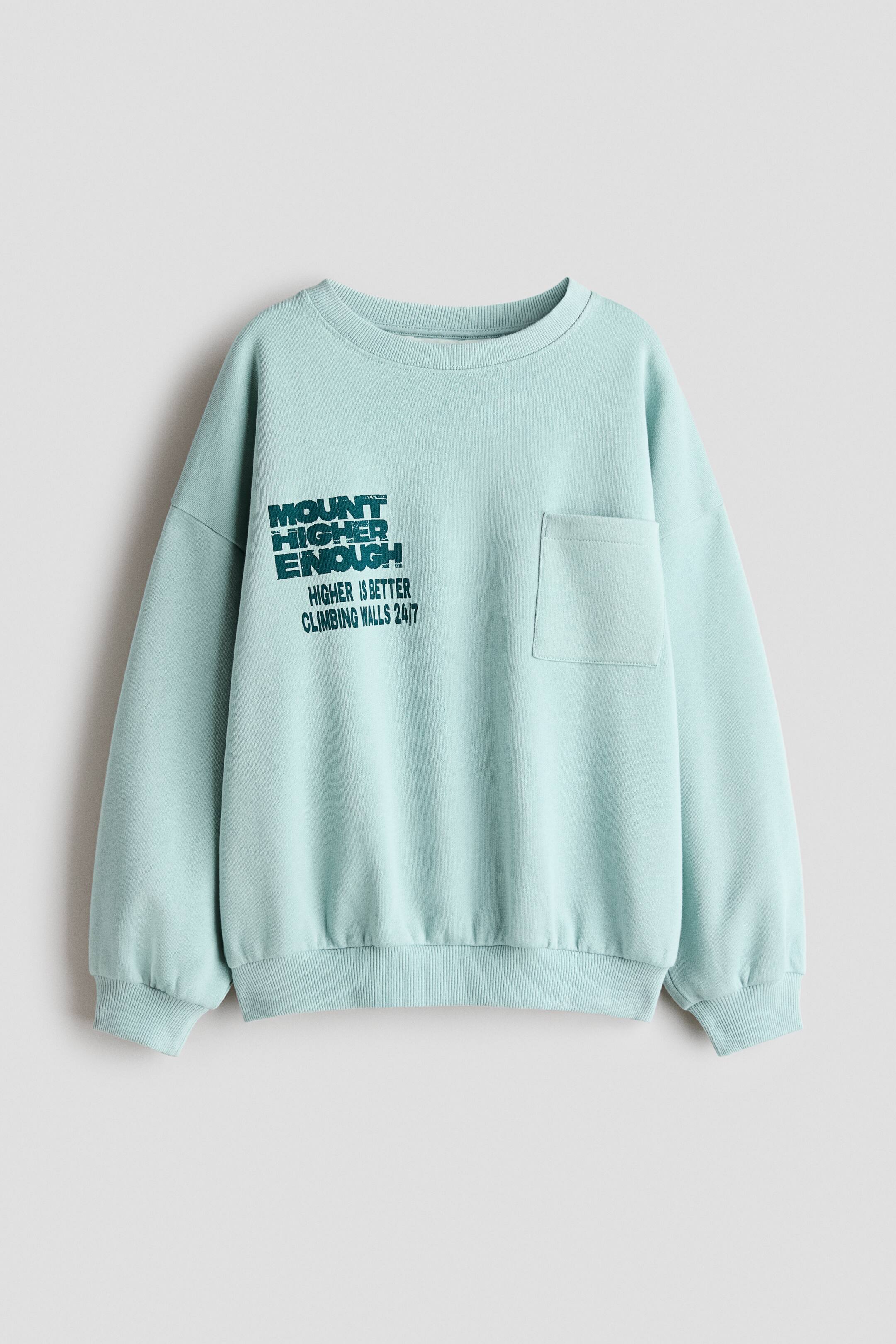 Agrandir l'image: Un sweat-shirt vert menthe clair est présenté de face, avec un col rond, des épaules tombantes, des poignets côtelés et un ourlet côtelé. Il présente une poche plaquée sur la poitrine gauche et un motif textuel effet vieilli vert foncé sur la poitrine droite.