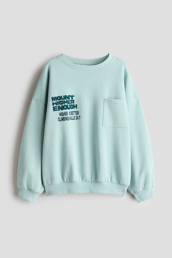 Un sweat-shirt vert menthe clair est prÃ©sentÃ© de face, avec un col rond, des Ã©paules tombantes, des poignets cÃ´telÃ©s et un ourlet cÃ´telÃ©. Il prÃ©sente une poche plaquÃ©e sur la poitrine gauche et un motif textuel effet vieilli vert foncÃ© sur la poitrine droite. - 1