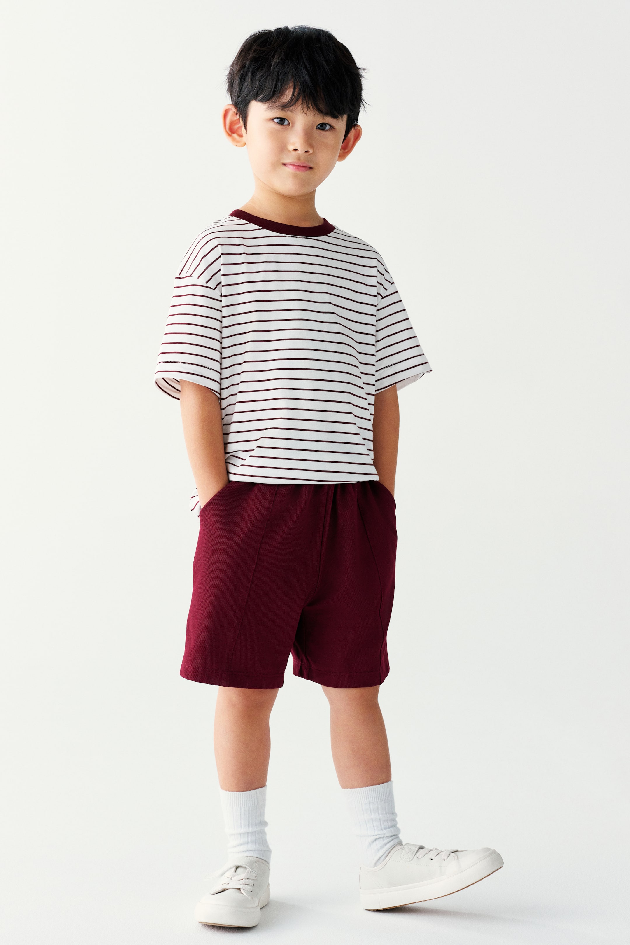 View larger image: The model is wearing a short-sleeved t-shirt and shorts set. The white crew-neck t-shirt features thin burgundy horizontal stripes and a solid burgundy collar, while the matching burgundy shorts have front seam details and side pockets where the model is resting their hands.