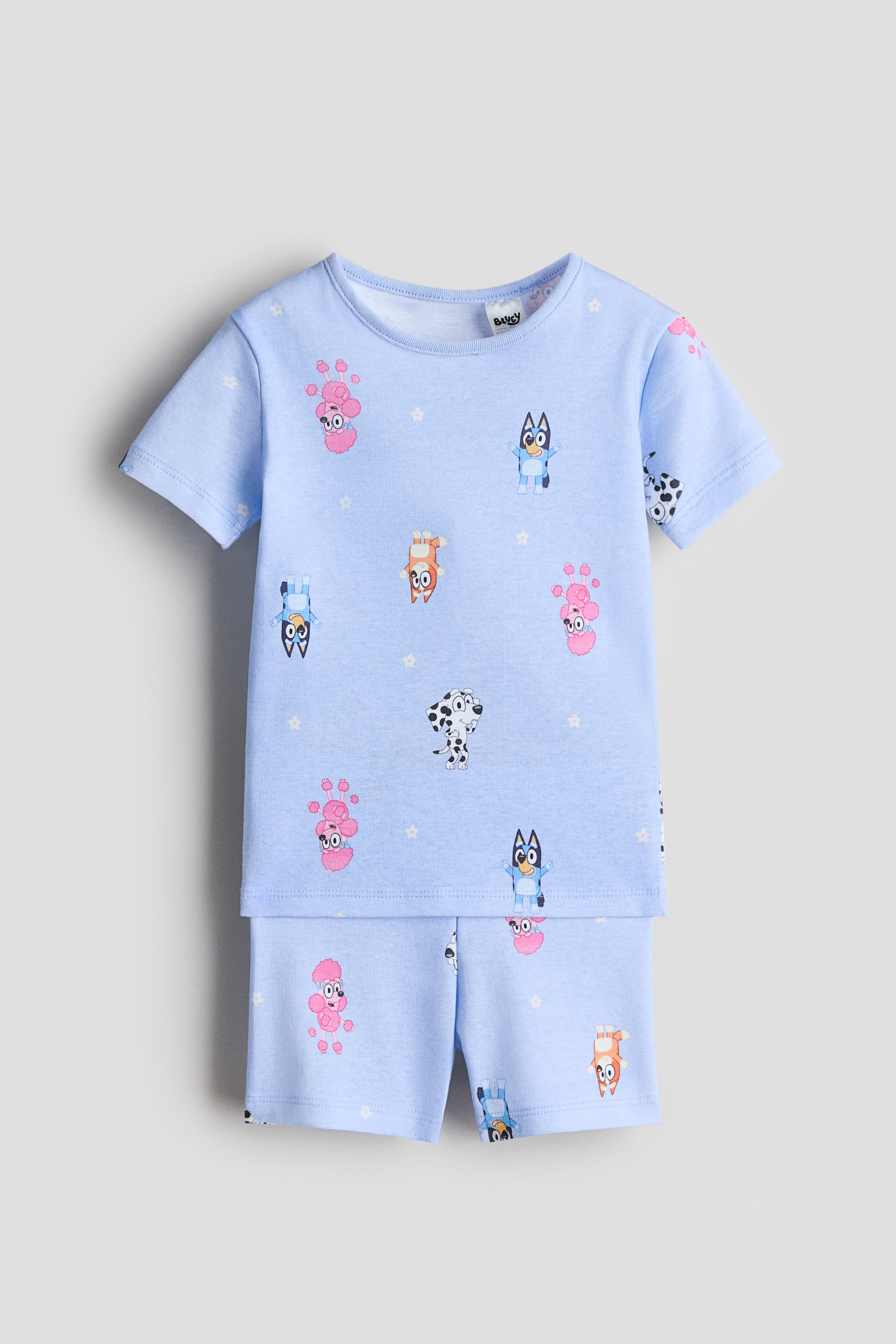 View larger image: A light blue pajama set, shown facing forward, features a short-sleeve top with a round, rib-trimmed neckline and matching shorts with a simple waistband. Both pieces display a repeating print of various cartoon dog characters and small white stars.