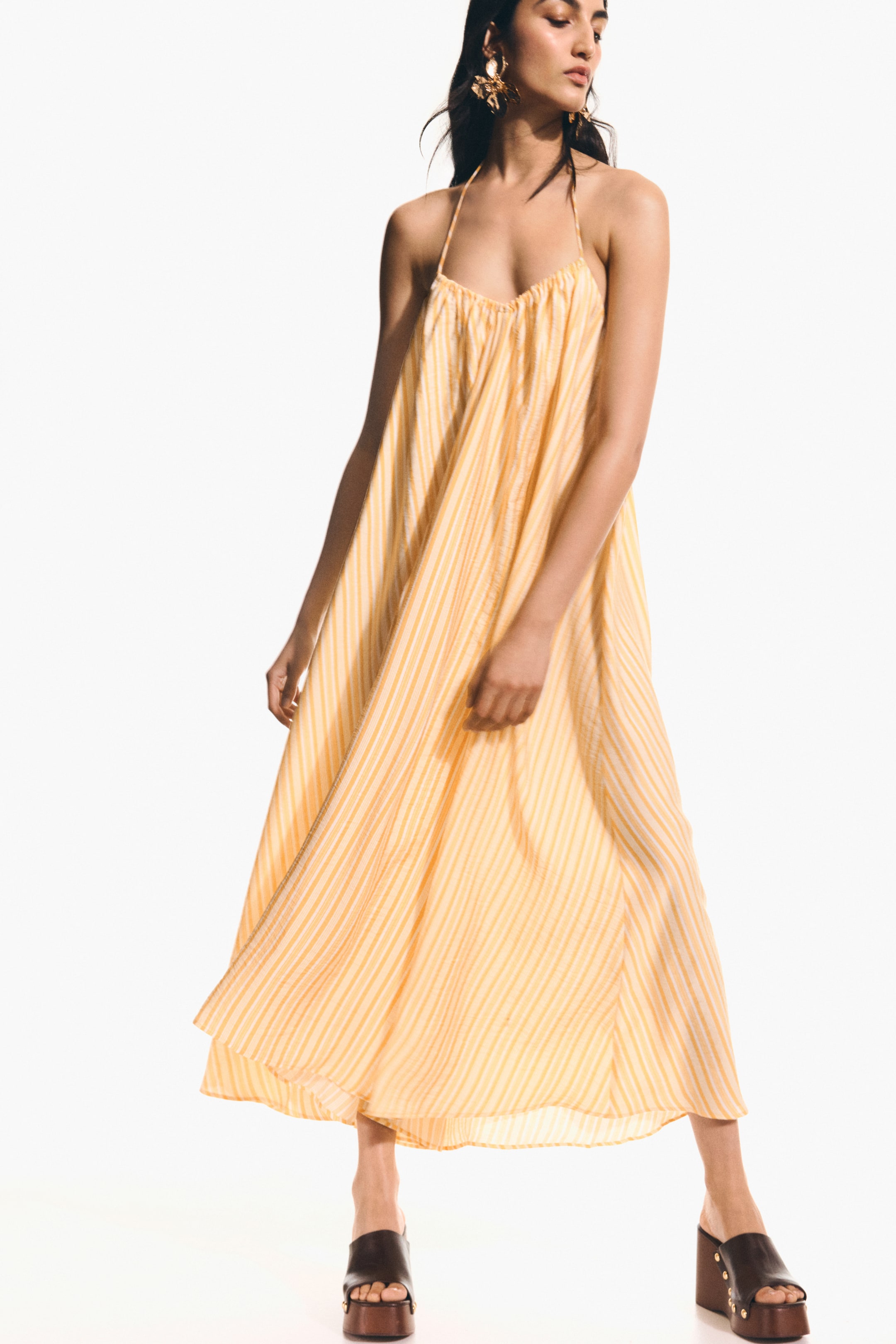 View larger image: A yellow maxi halter neck dress featuring thin vertical white stripes. It showcases a gathered bust and a flowing A-line silhouette, reaching the ankles.