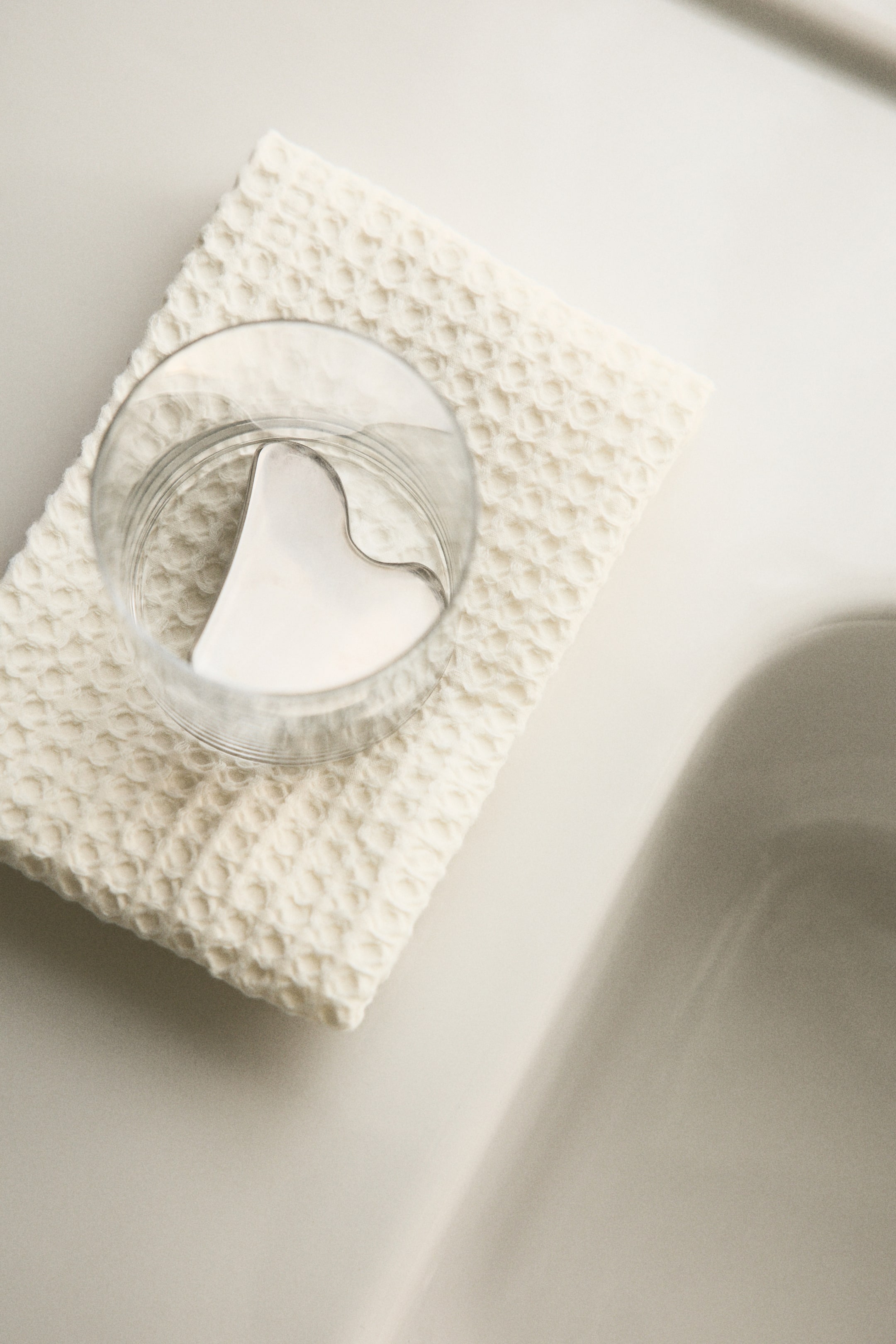 Voir une image plus grande: Une serviette pour invités de couleur blanc cassé présente une texture unique en nid d'abeille. Un verre transparent repose sur la serviette, contenant un outil gua sha en forme de cœur de couleur claire.