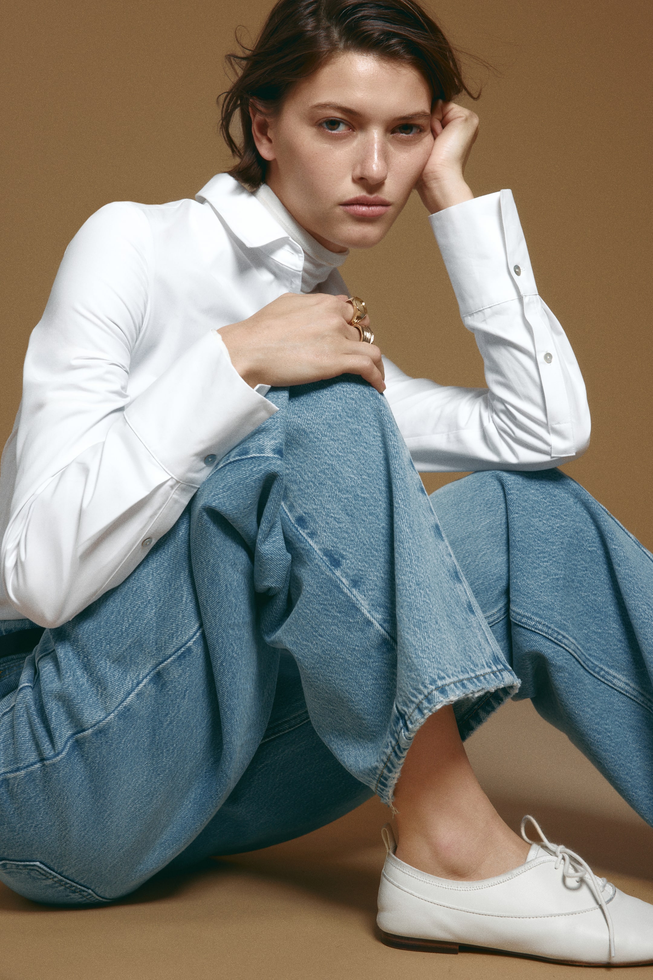 Se større billede: Blå denimjeans med en buet, voluminøs ben-silhuet bæres af modellen, der sidder på gulvet. Jeansen er stylet med en hvid skjorte med knapper og lange ærmer, lagt over en rullekrave, og kombineret med hvide flade snøresko.