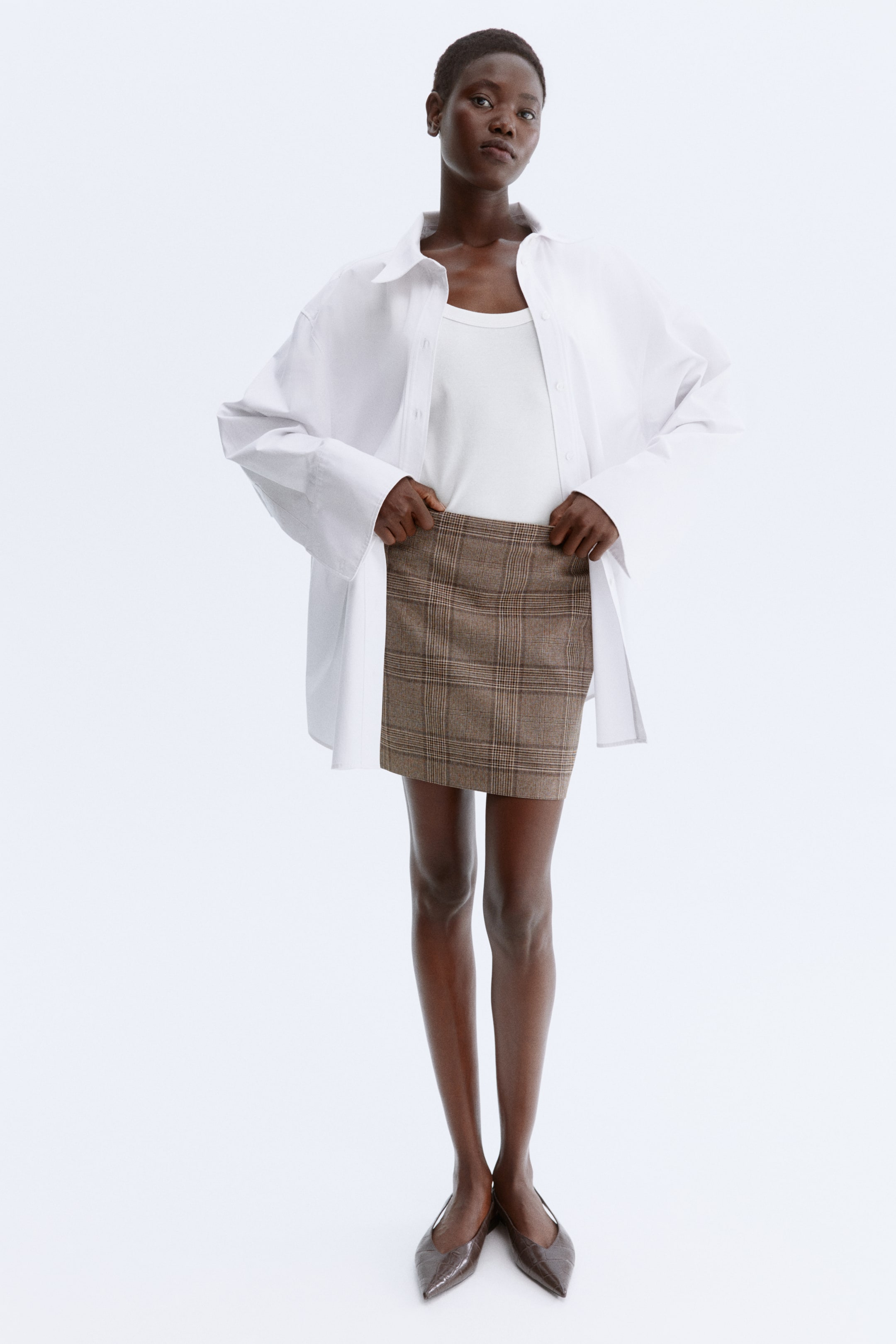 View larger image: A brown plaid mini skirt is being presented from a front-facing angle, with hands holding its waistband. The skirt is featuring a classic plaid pattern with lighter brown and cream lines, and showcasing a straight, short cut that ends well above the knee.