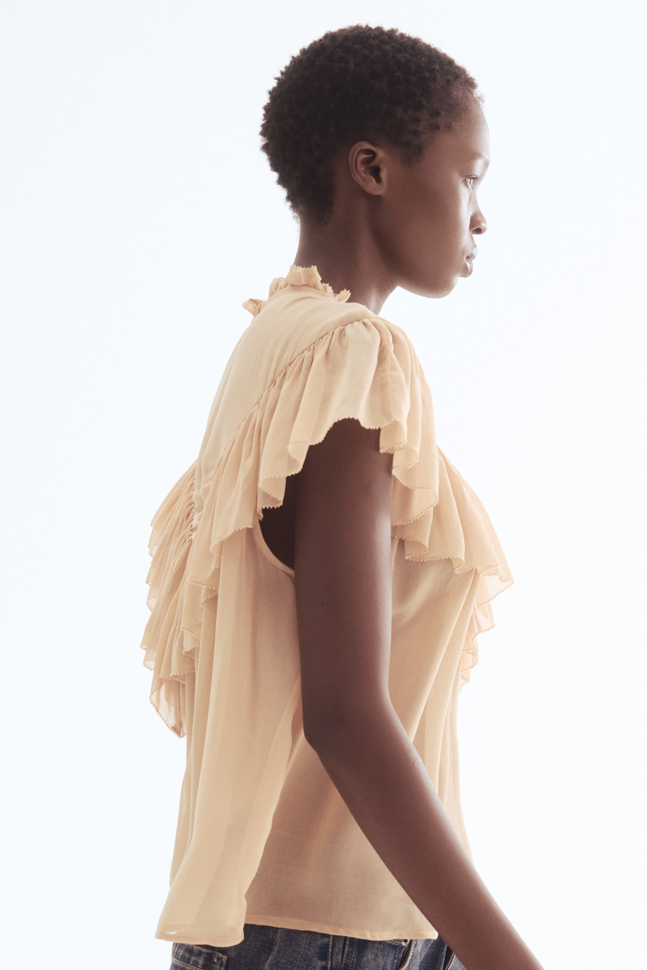 View larger image: Viewed from a side profile facing right, a light peach sleeveless top is featuring a ruffled high neck and tiered ruffles cascading from the shoulders down the chest and back, with its hem resting over blue denim jeans.