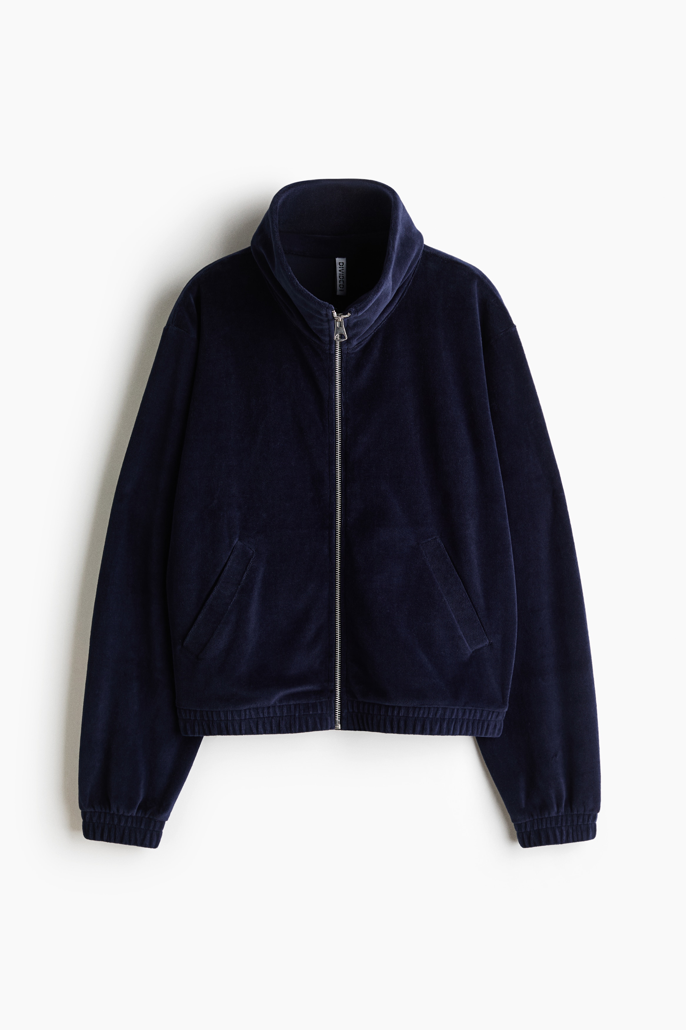 A dark blue velour track jacket featuring a double-layered stand collar, front zipper, and dropped shoulders is being worn by the model. The model is holding the elasticated hem of the jacket, which is styled with matching wide-leg velour trousers.