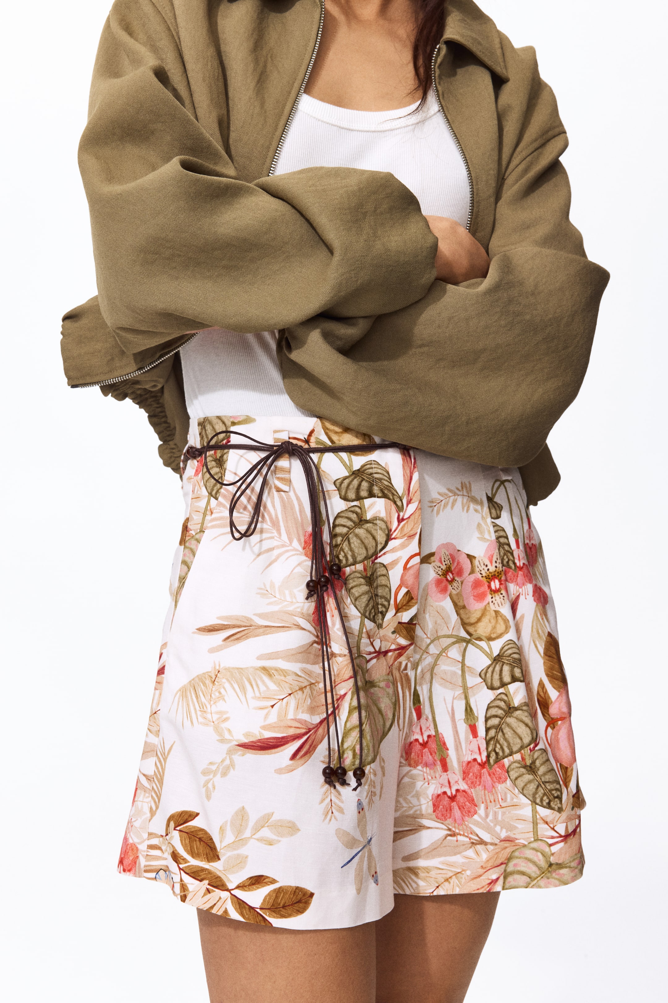 이미지 확대 보기: 흰색 반바지는 식물 무늬를 보여주며, 연한 갈색 잎, 분홍색 꽃, 파란색 잠자리가 특징입니다. 허리에는 작은 나무 구슬이 달린 짙은 갈색 끈이 묶여 있으며, 반바지는 여유로운 핏으로 허벅지 중간까지 내려옵니다.