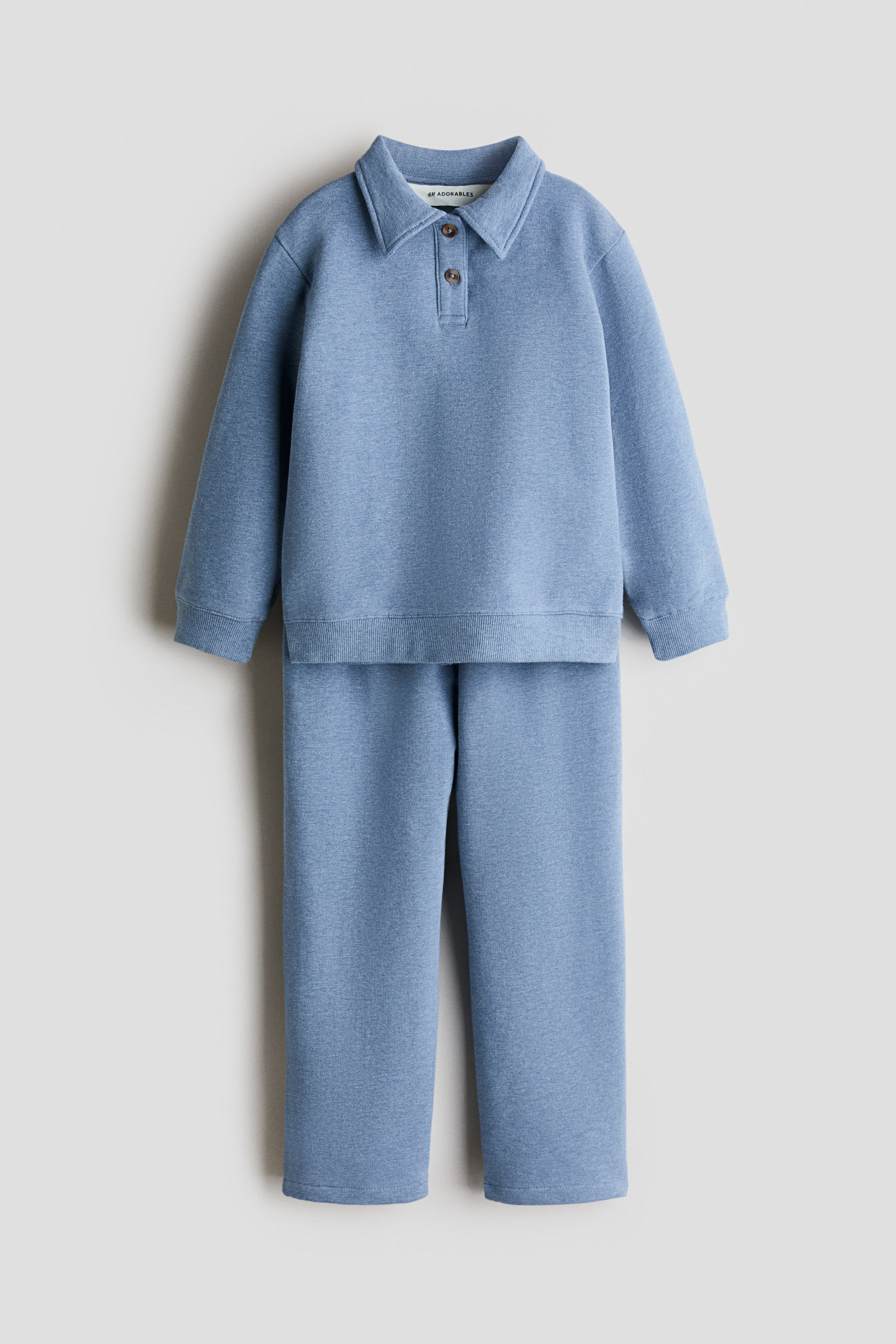 Visualizza l'immagine ingrandita: Un completo due pezzi blu-grigio, realizzato in morbido tessuto felpato, presenta un top a maniche lunghe con colletto a revers, abbottonatura a due bottoni, spalle scese e polsini e fondo a costine. I joggers abbinati presentano un elastico in vita e gambe dritte.