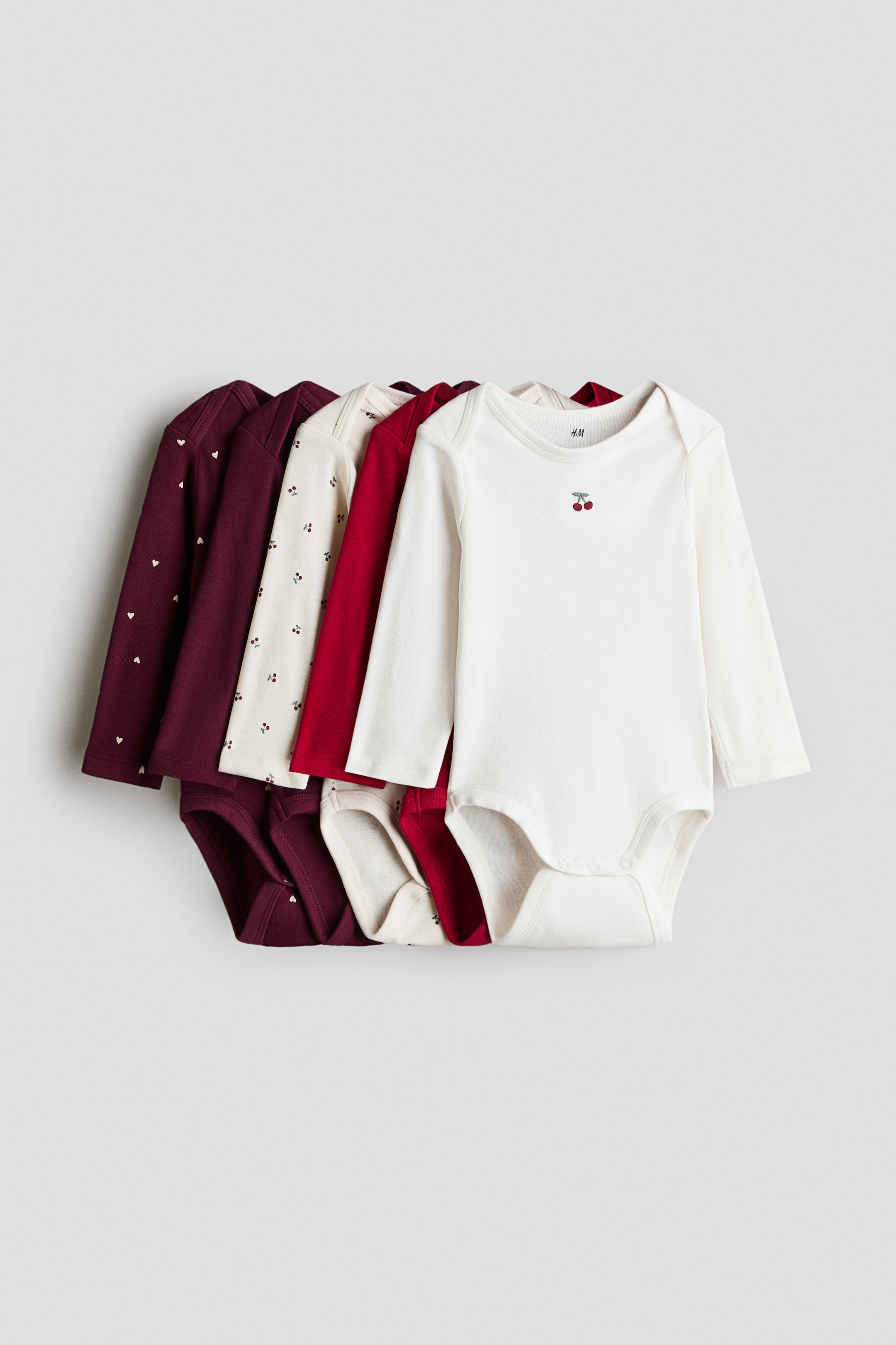 View larger image: Five long-sleeve cotton jersey bodysuits are overlapping in a front-facing view, showcasing their envelope necklines and press-stud closures at the bottom. The collection features a mix of solid and patterned designs in shades of off-white, red, and dark burgundy.