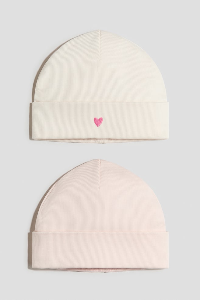 Dois gorros exibem a sua vista FRONTAL, feitos em jersey de algodão macio com textura canelada e barras viradas. O gorro superior é de cor creme e apresenta um pequeno coração rosa bordado, enquanto o gorro inferior é rosa claro. - 1