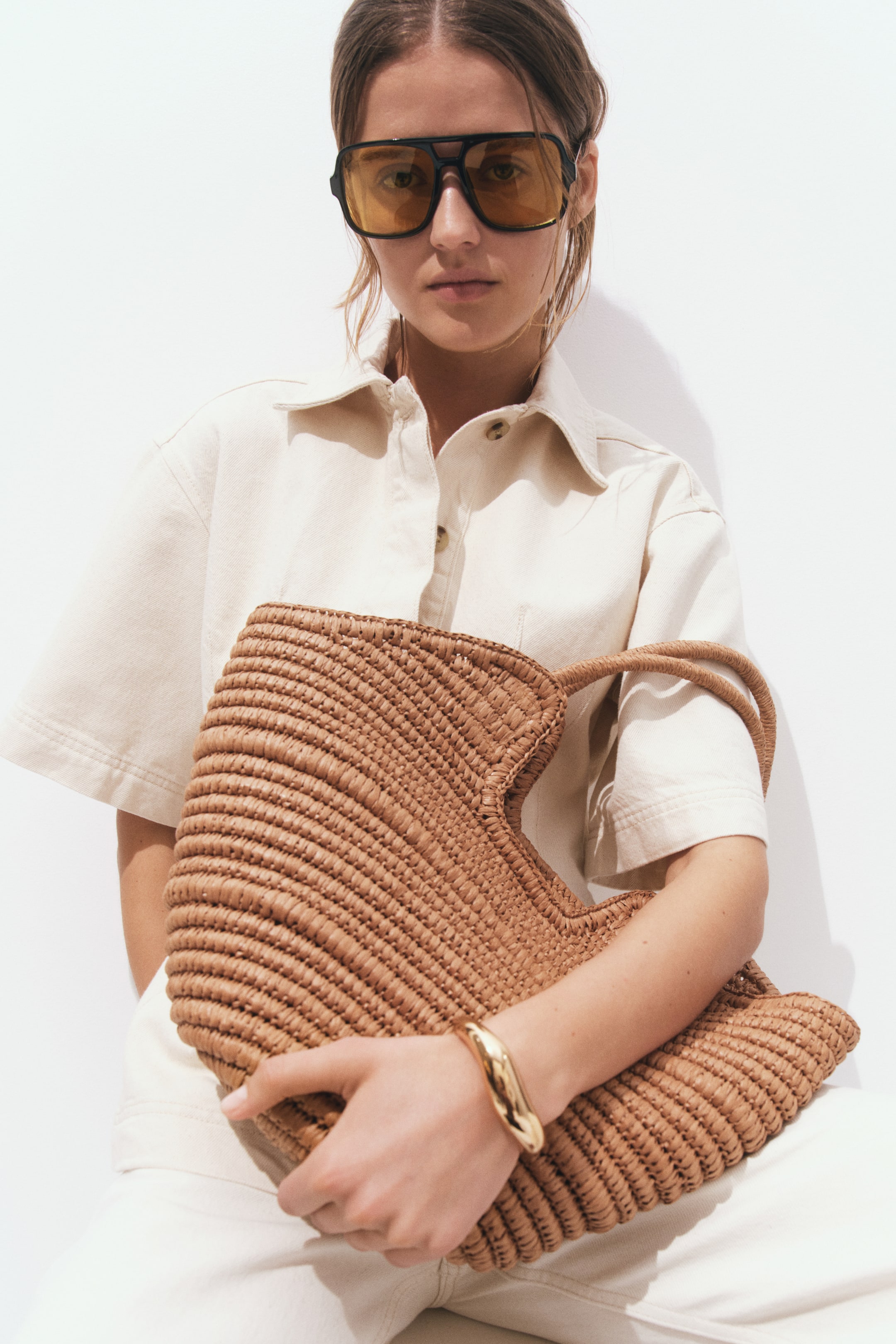 View larger image: A natural brown raffia-like tote bag is being held, showcasing a unique horizontal weave pattern across its body and two integrated top handles. Its wide, rounded top opening is facing forward.