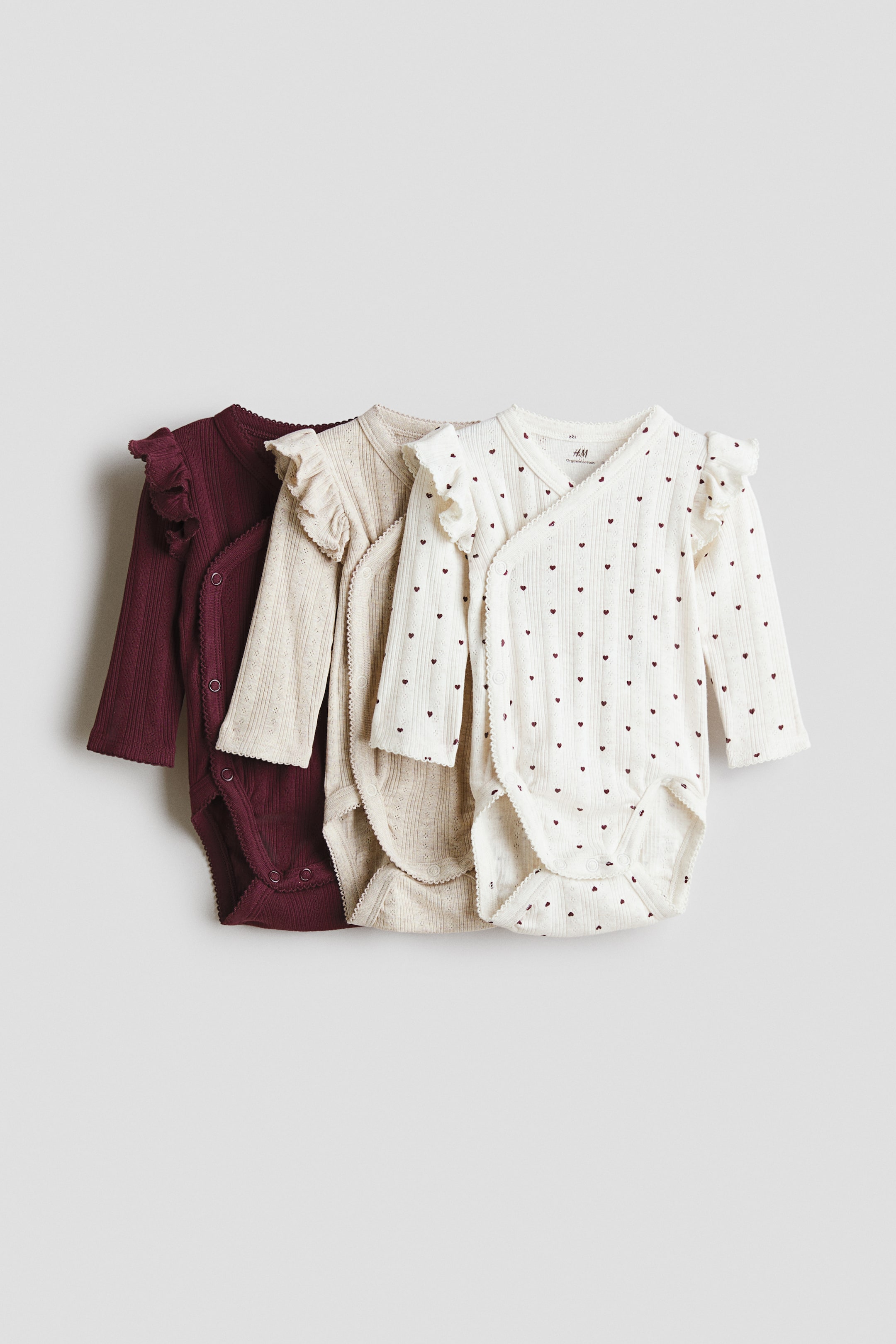 View larger image: Three long-sleeve baby bodysuits in soft pointelle jersey are overlapped and facing front, showcasing a wrap design with press studs, delicate picot trims, and ruffled shoulder details. The set includes a solid dark burgundy bodysuit, a solid light beige bodysuit, and an off-white bodysuit featuring an all-over small red heart pattern.