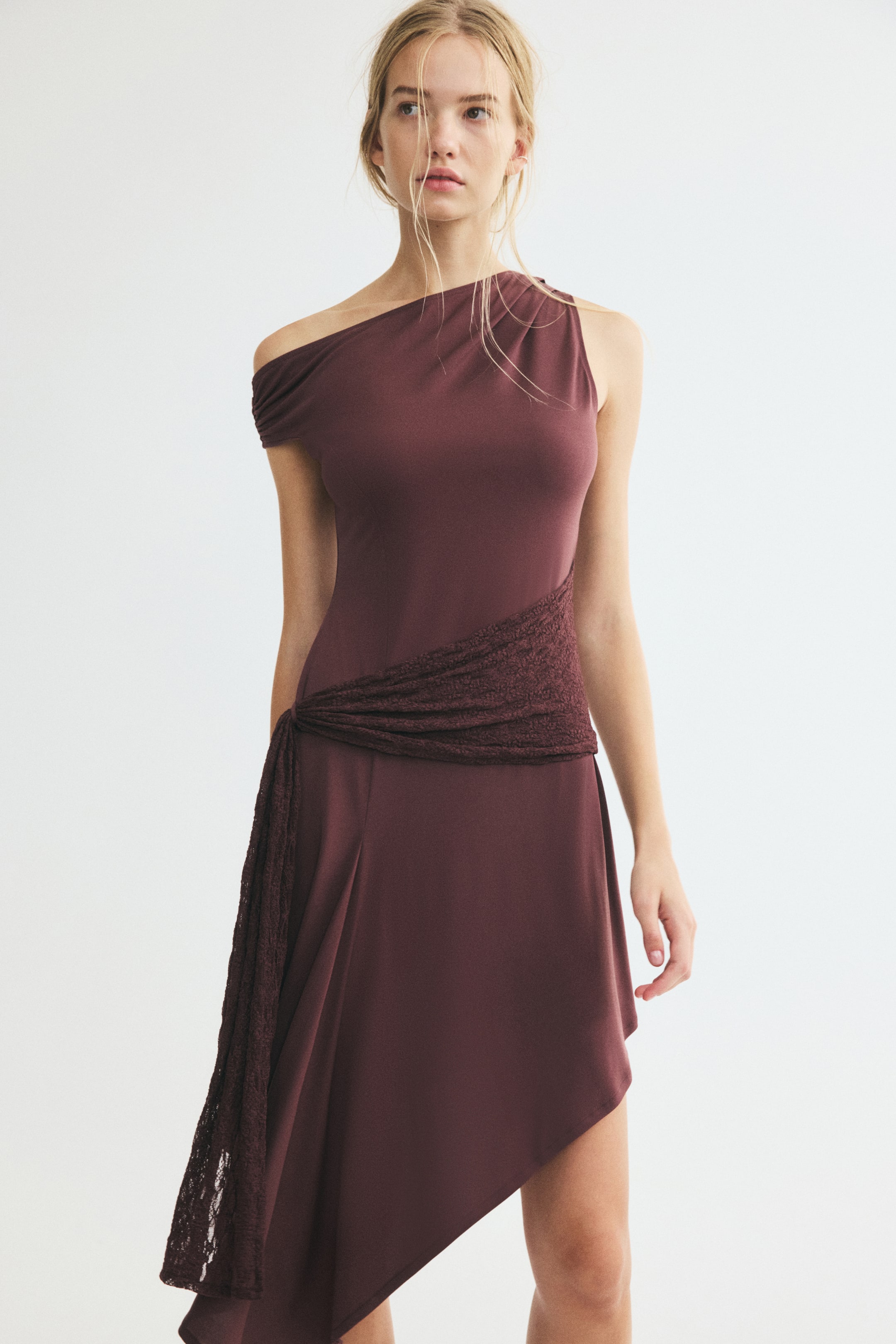 View larger image: A draped lace panel dress in a deep plum hue is being worn by the model, featuring an asymmetrical one-shoulder neckline and a fitted bodice. The dress is detailed with a textured lace panel wrapped around the waist that drapes down the side, ending in an asymmetrical hemline.