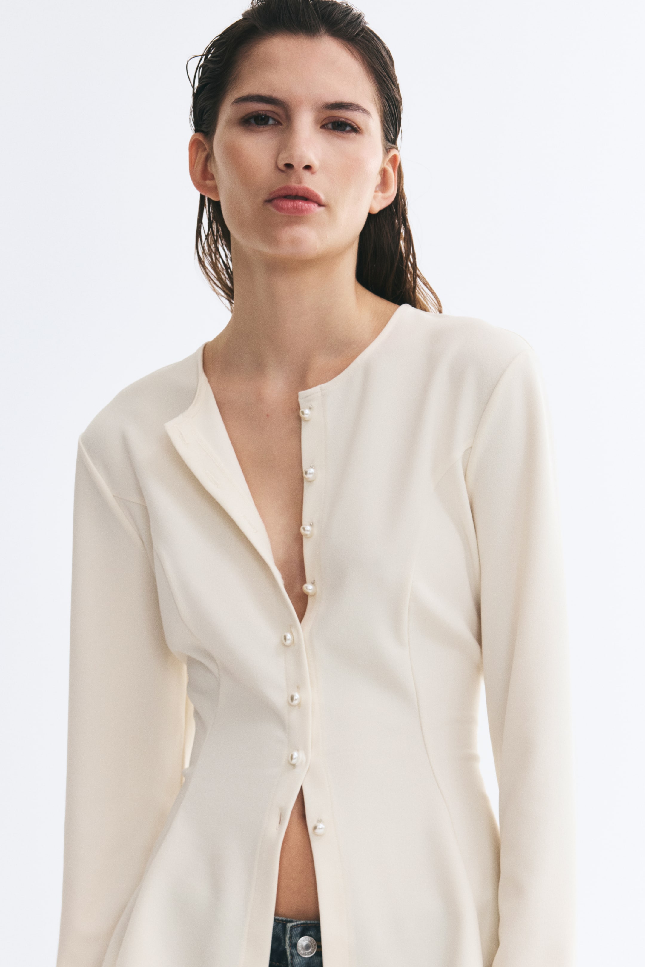 View larger image: A cream-coloured, long-sleeved top featuring a round neckline and a front closure with small pearl buttons is worn by the model. The top is styled partially unbuttoned at the neck and hem, revealing the waistband of blue denim jeans.