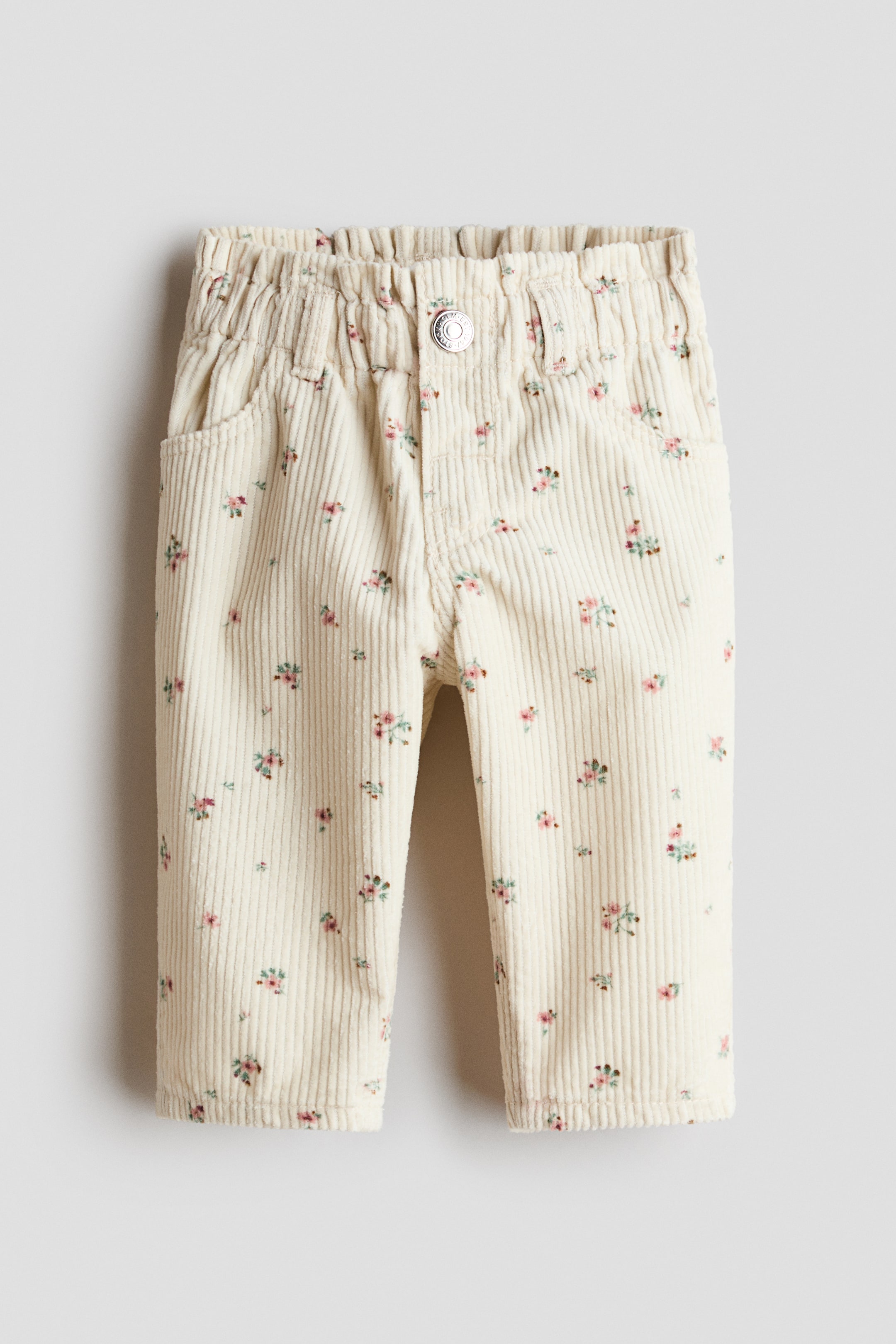 Agrandir l'image: Pantalon en velours côtelé de couleur crème, orienté vers l'avant, avec un imprimé intégral de petits motifs floraux roses et verts. Le pantalon présente une taille élastiquée, une fermeture à bouton argenté unique et deux poches plaquées arrondies à l'avant.