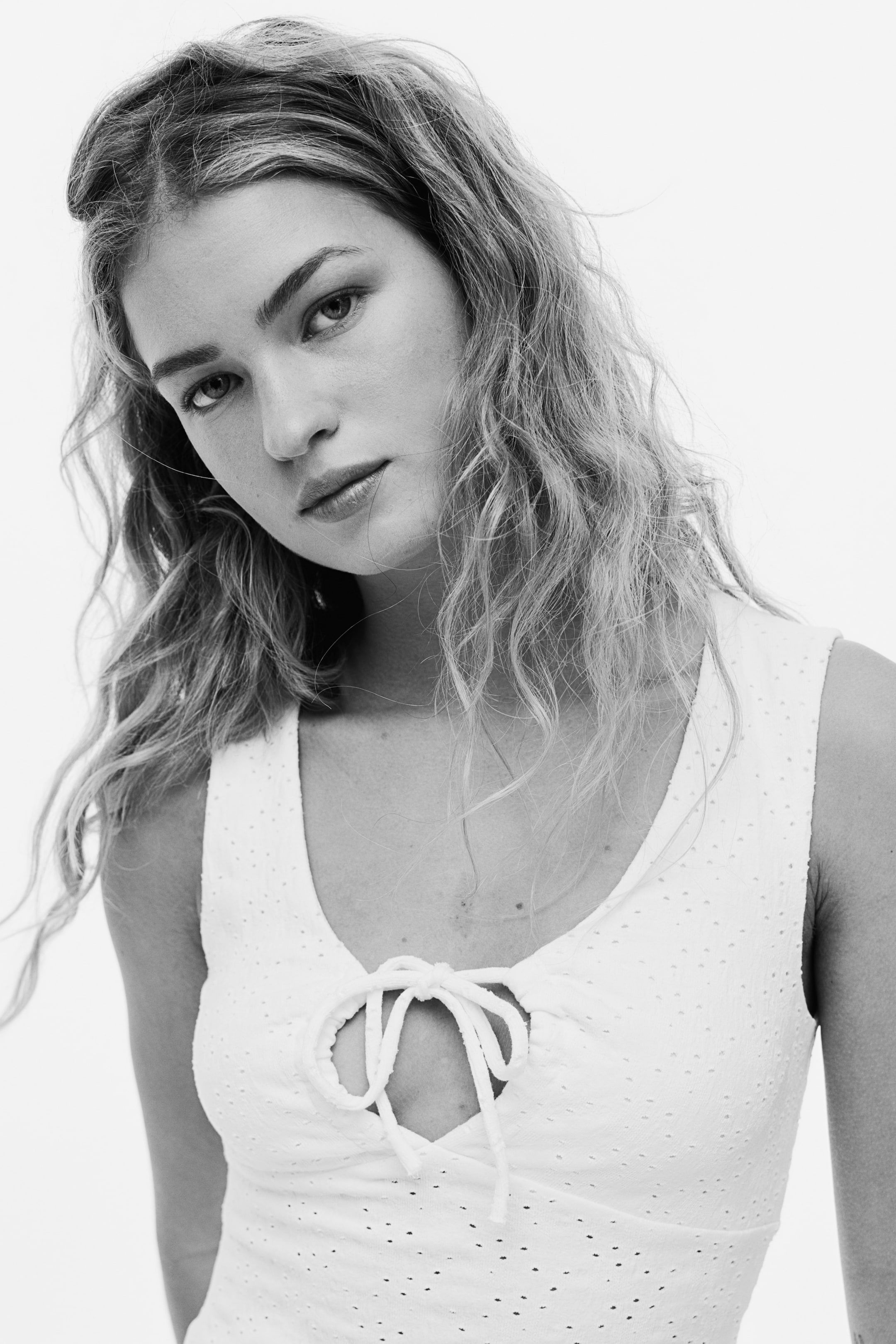 View larger image: A white sleeveless top showcases a V-neckline with a keyhole cutout at the centre front. The cutout is tied with a bow, and the fabric displays an all-over eyelet pattern.
