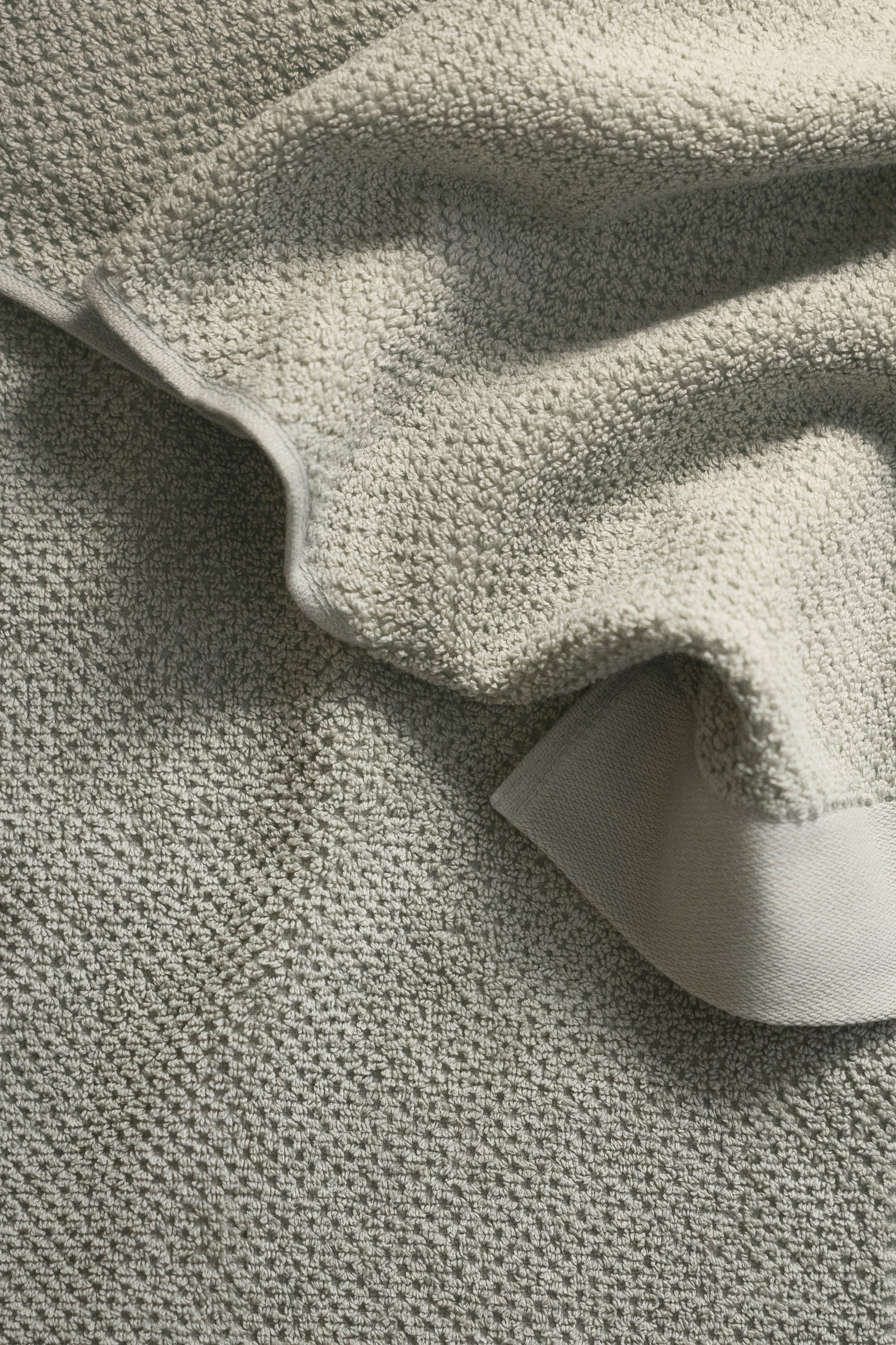 View larger image: A light sage-green textile presents two distinct textures. The primary surface features a raised waffle weave, while a folded section reveals a softer, looped terry texture with a smooth, flat edge.