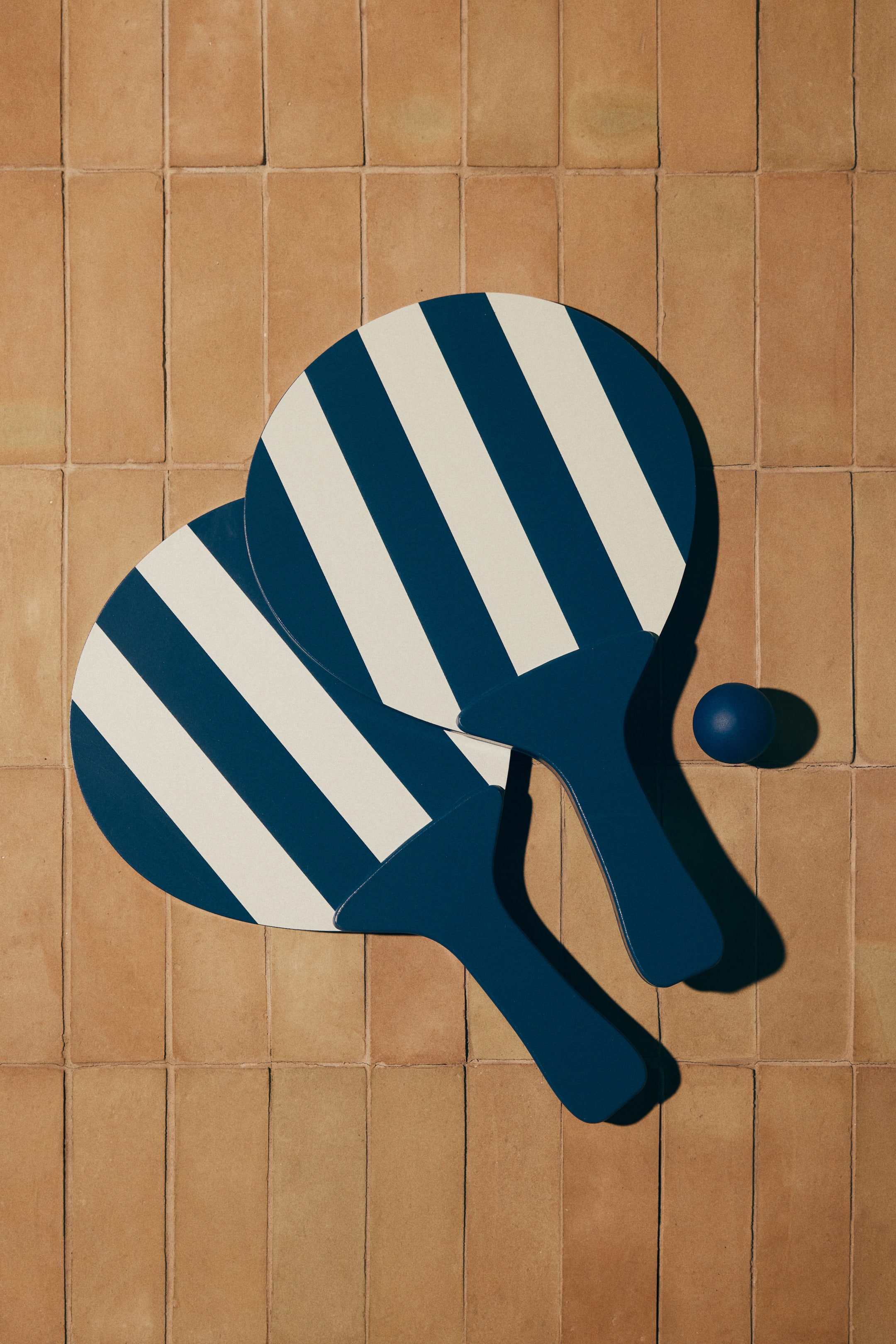 View larger image: Two beach paddles are overlapping, showcasing diagonal navy blue and off-white stripes on their circular heads and solid navy blue handles. A small, solid navy blue ball is positioned next to the paddles.