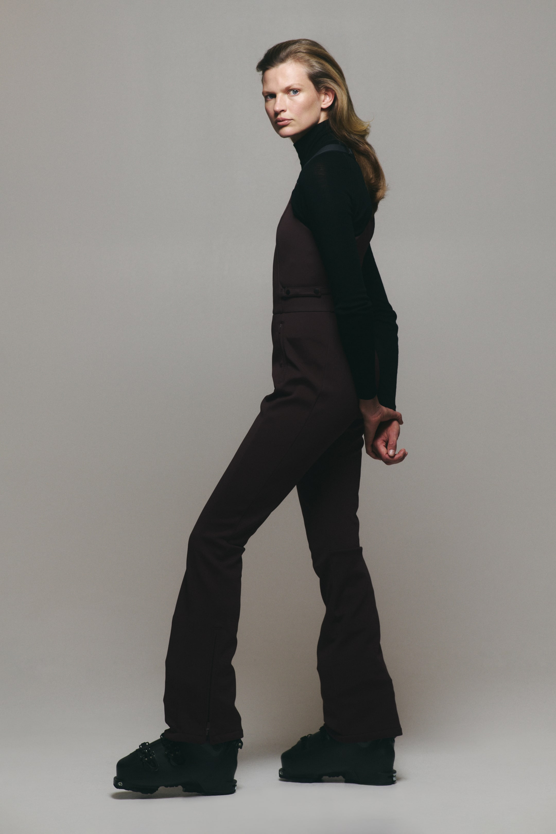 View larger image: The model is wearing dark brown ski overalls, layered over a black long-sleeved turtleneck sweater. The slim-fitting overalls feature a high-waisted bib, side-button detailing, and bootcut legs resting over black ski boots.