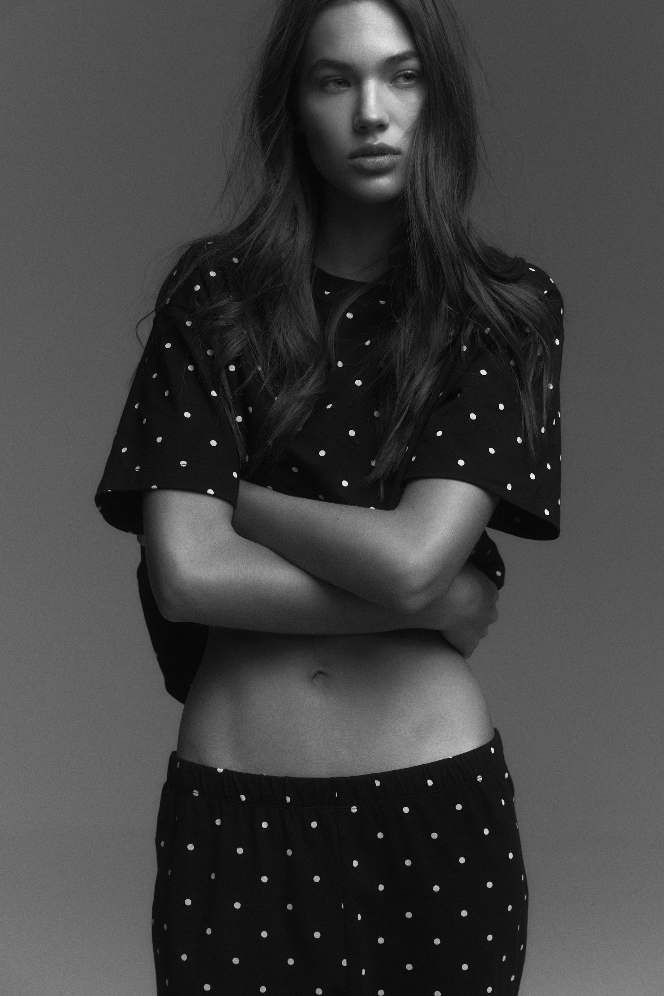 View larger image: Pyjamas, featuring a black T-shirt with a round neckline, dropped shoulders, and white polka dots, are being worn by the model. The T-shirt is being worn cropped, revealing the midriff, and the matching bottoms are showcasing an elasticated waist as the model is crossing her arms over her torso.