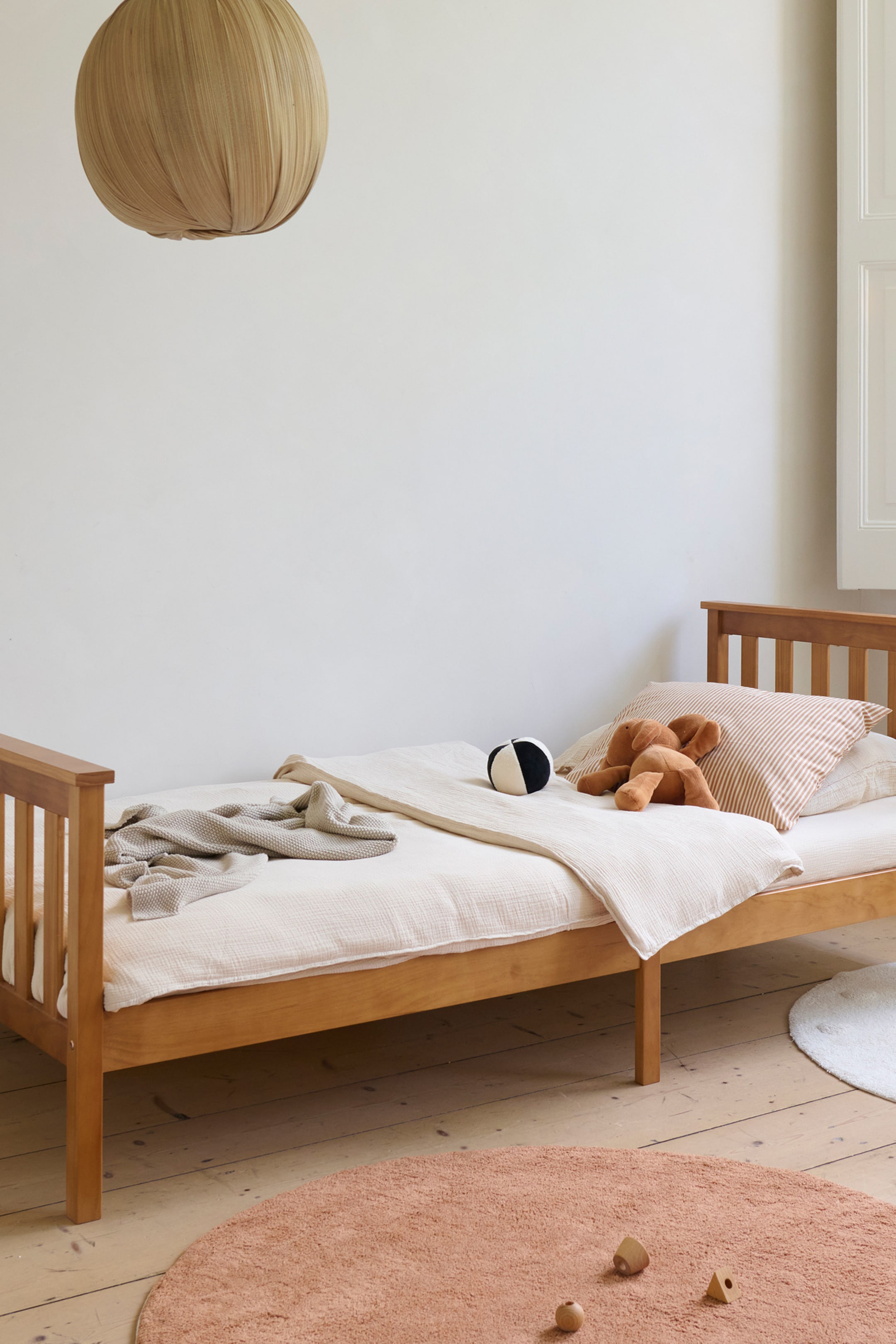 Grotere afbeelding bekijken: Een bedframe van natuurlijk hout, met een latten hoofdbord en voetbord, wordt onder een hoek gepresenteerd, waarbij de volledige lengte zichtbaar is. Het is voorzien van licht crème beddengoed, met een lichtgrijze wafelgebreide deken over het voeteneinde gedrapeerd, en een gestreept kussen, een bruine knuffel en een zwart-witte bal die op het hoofdeinde rusten.